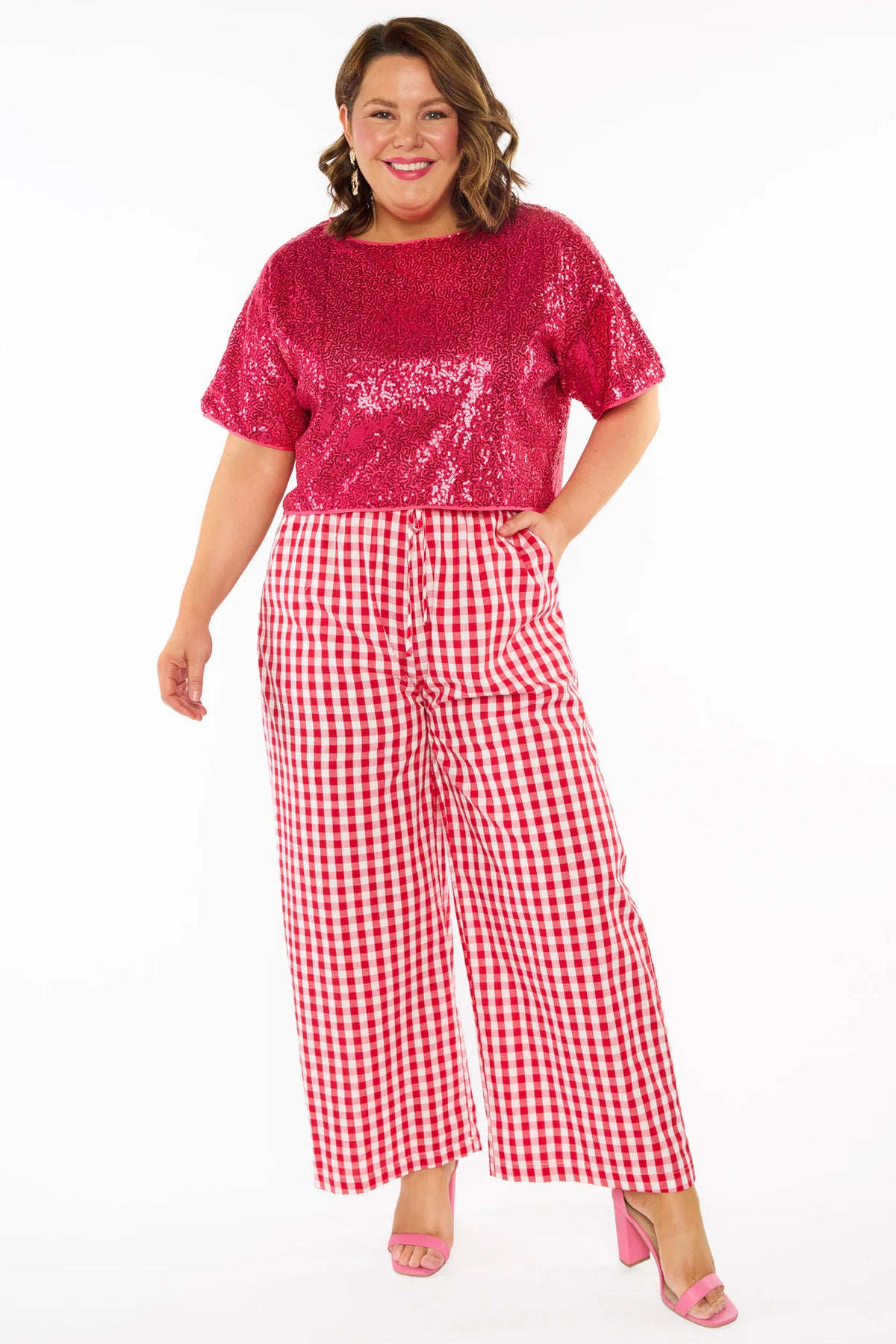 A woman modelling a two-piece outfit consisting of a bright pink sequins short-sleeve top and red-and-white gingham wide-leg pants, paired with pink heels, against a plain white background.