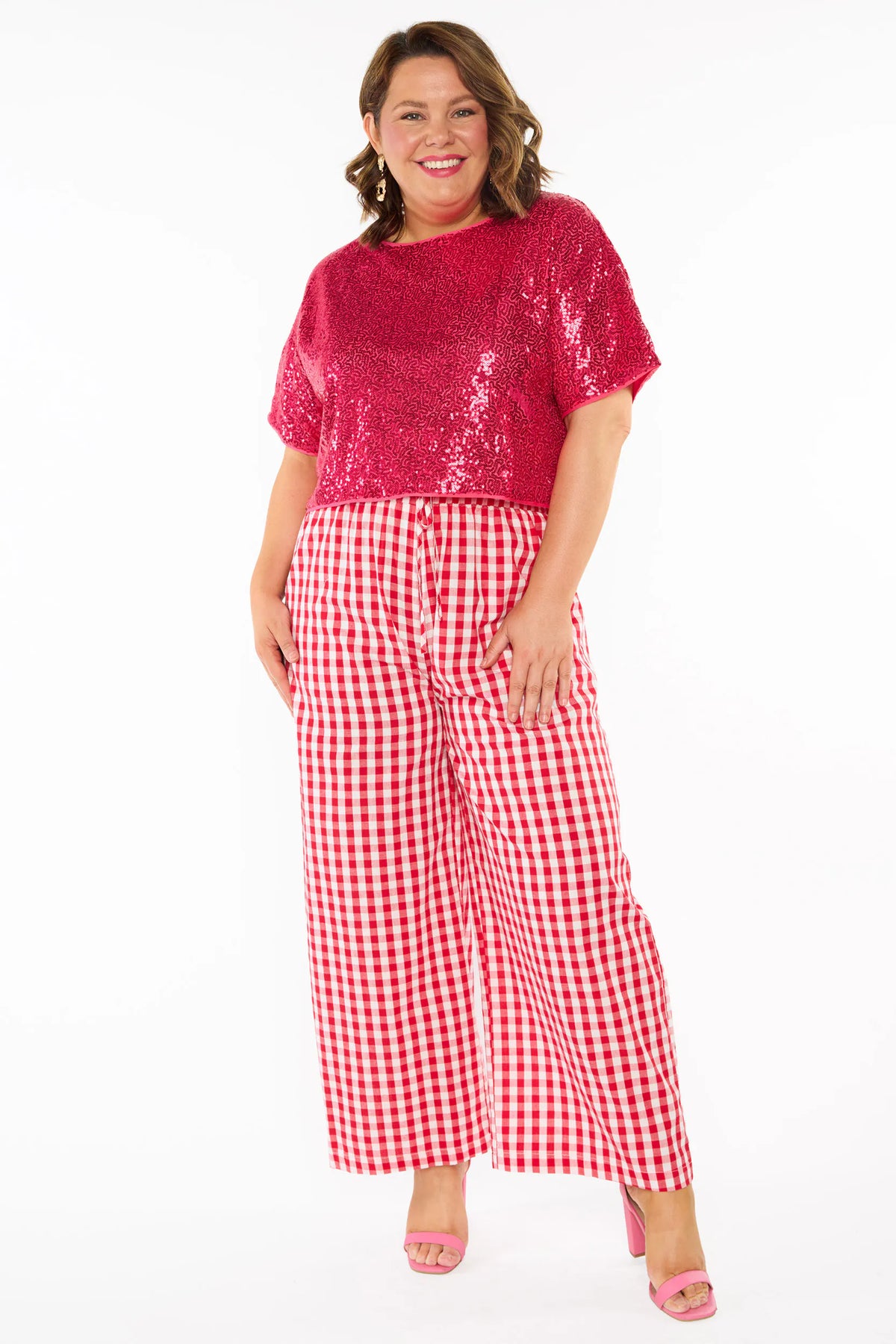 A woman modelling a two-piece outfit consisting of a bright pink sequins short-sleeve top and red-and-white gingham wide-leg pants, paired with pink heels, against a plain white background.