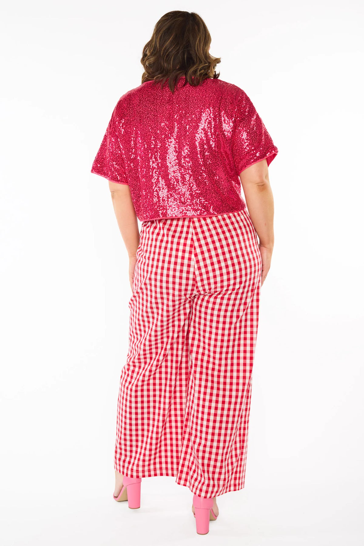 Back view of a woman modelling a two-piece outfit consisting of a bright pink sequins short-sleeve top and red-and-white gingham wide-leg pants, paired with pink heels, against a plain white background.