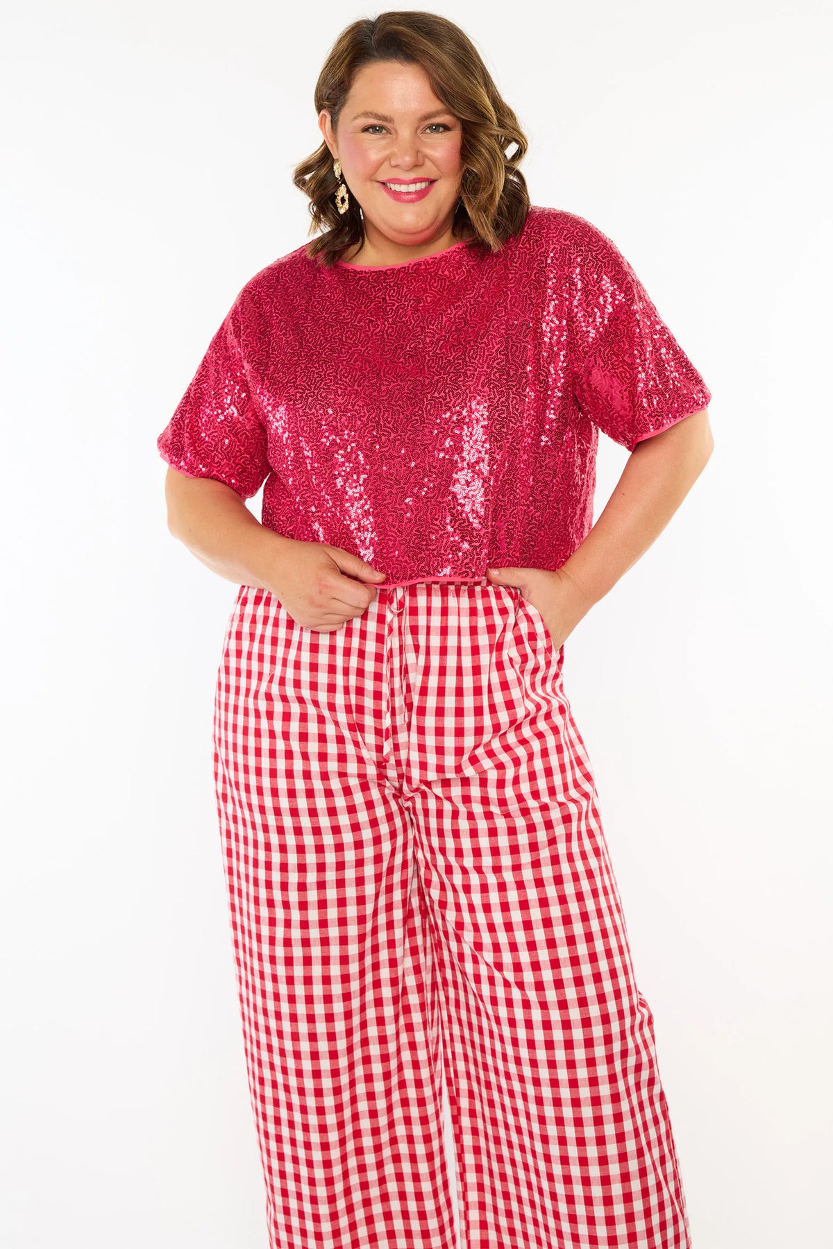A woman modelling a two-piece outfit consisting of a bright pink sequins short-sleeve top and red-and-white gingham wide-leg pants, paired with pink heels, against a plain white background.