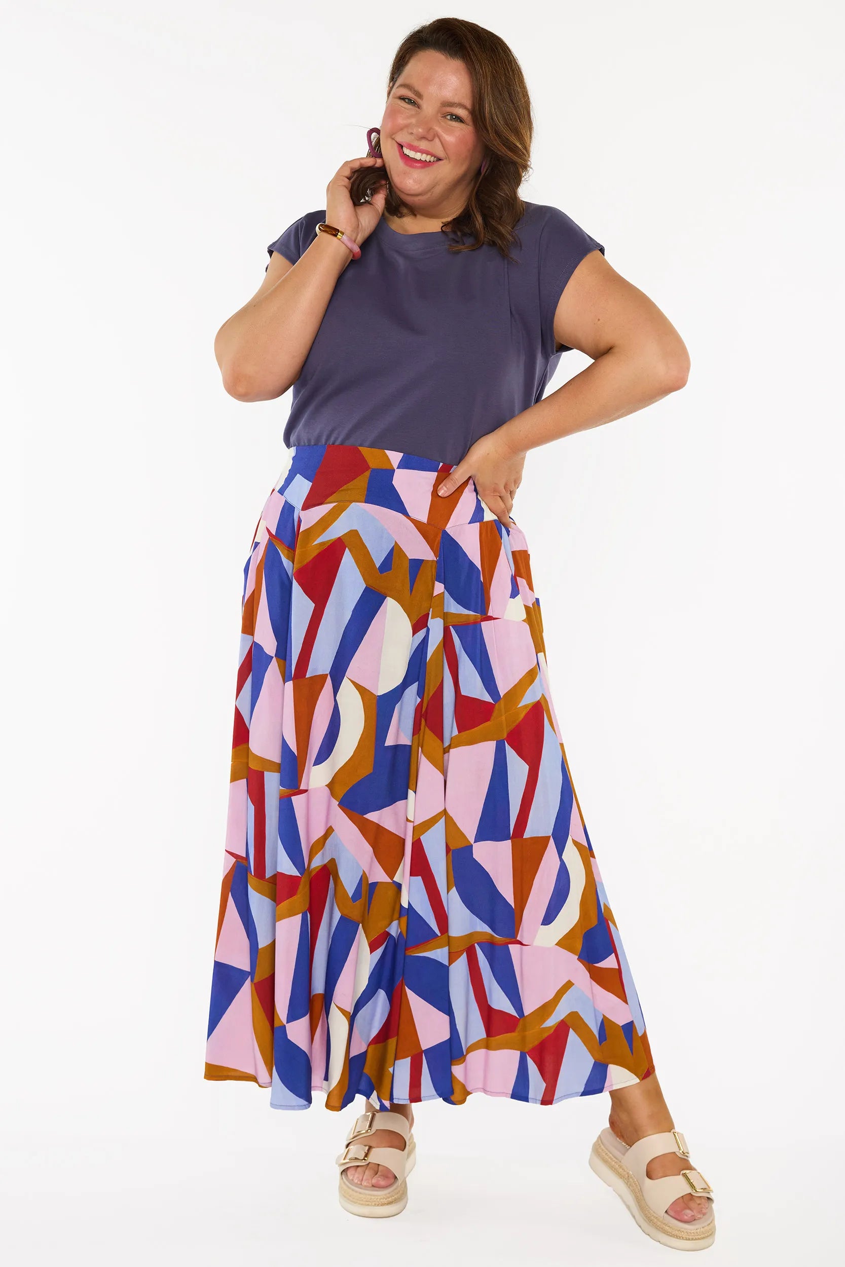 A woman modelling a sleeveless, high-waisted maxi skirt with a colourful geometric abstract print in blue, pink, red, and brown, paired with a solid navy blue top and beige platform sandals, against a plain white studio background.