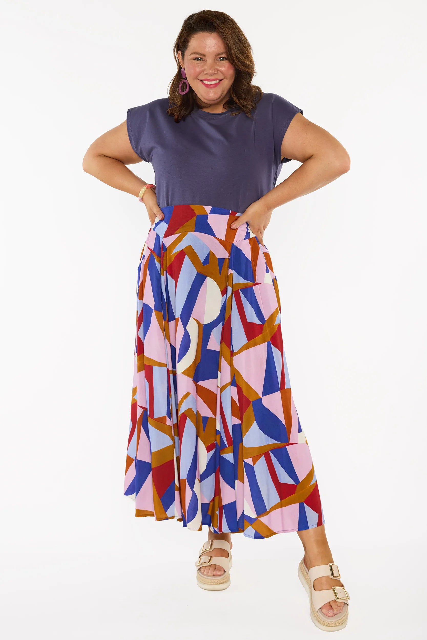 A woman modelling a sleeveless, high-waisted maxi skirt with a colourful geometric abstract print in blue, pink, red, and brown, paired with a solid navy blue top and beige platform sandals, against a plain white studio background.