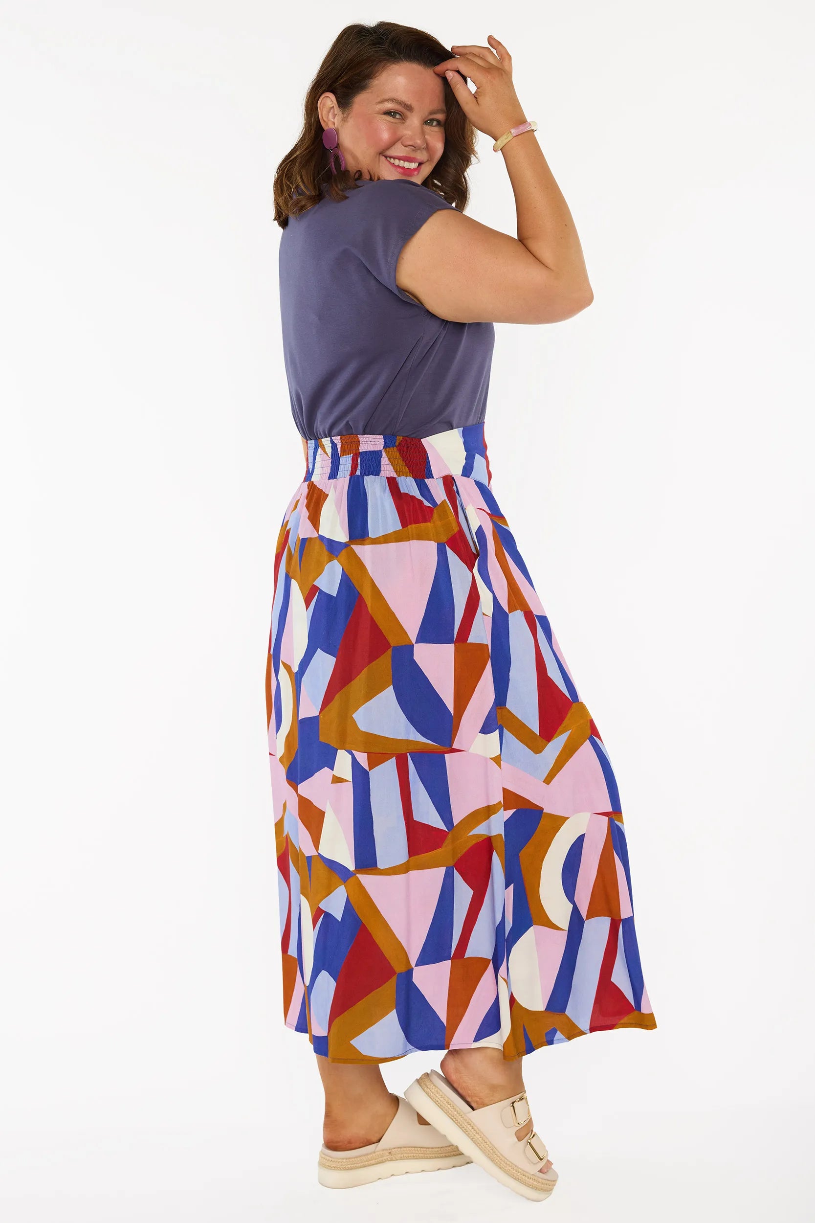 A woman modelling a sleeveless, high-waisted maxi skirt with a colourful geometric abstract print in blue, pink, red, and brown, paired with a solid navy blue top and beige platform sandals, against a plain white studio background.