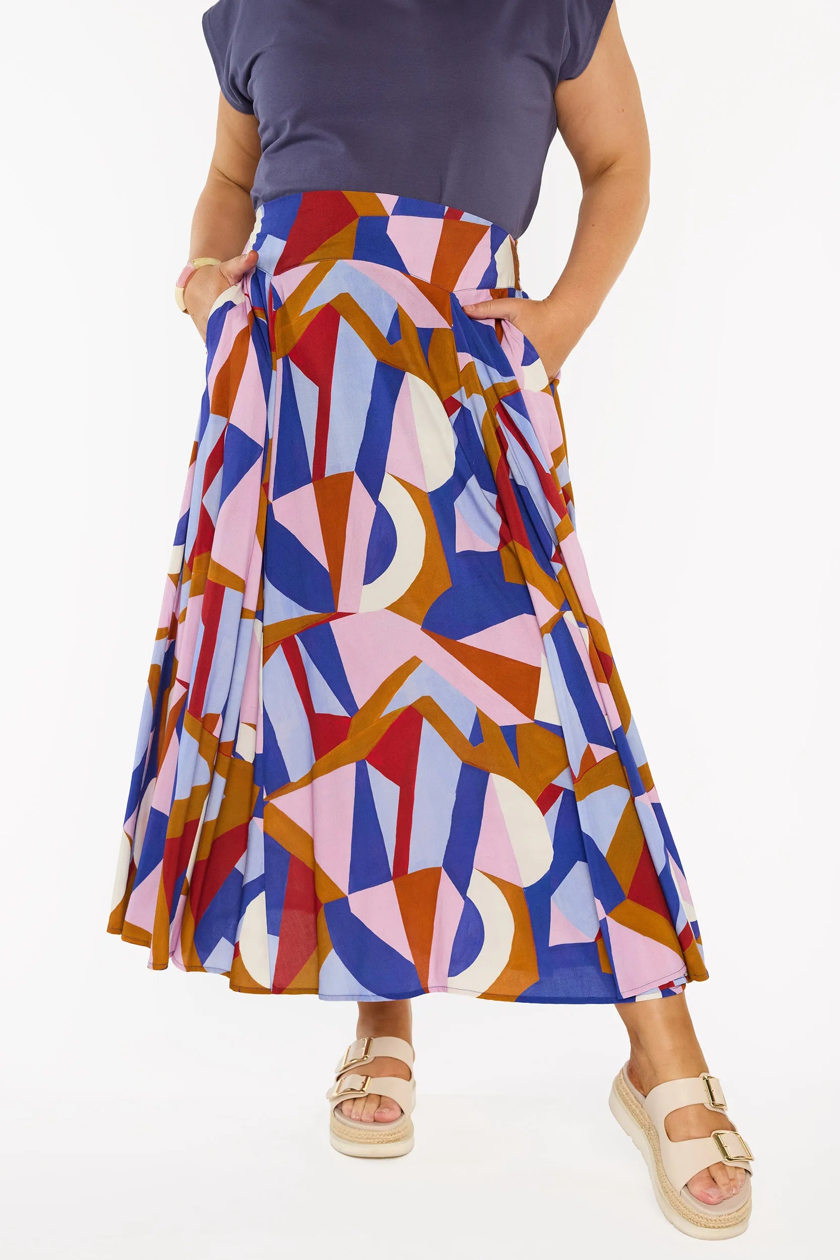 A model wearing a high-waisted, pleated midi skirt with a colourful geometric abstract print in blue, pink, red, and brown, styled with a navy blue top and beige platform sandals.