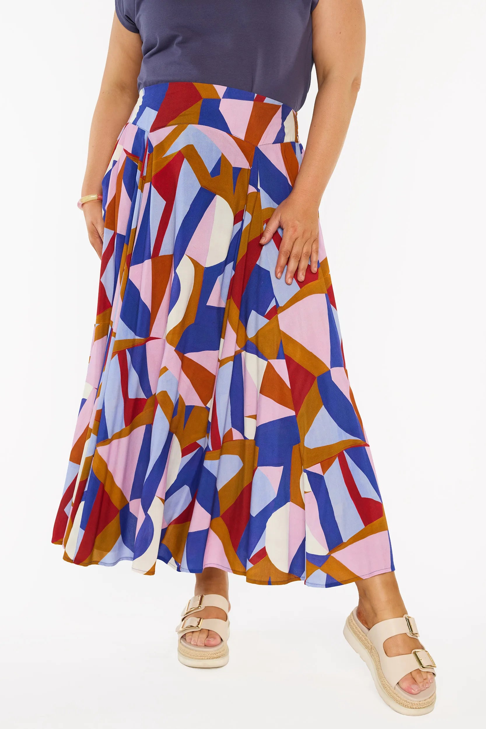 A model wearing a high-waisted, tiered maxi skirt with a colourful abstract geometric print in shades of blue, pink, red, orange, and white. The model is also wearing a navy blue top and beige platform sandals, photographed against a clean white background.