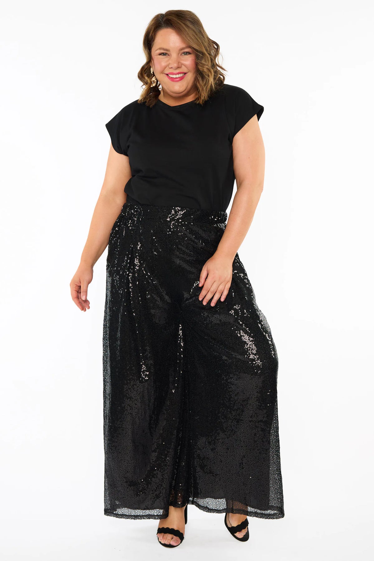 A woman modelling a black short sleeves top and a black sequin wide leg pant, standing against a plain white studio background with black heels.