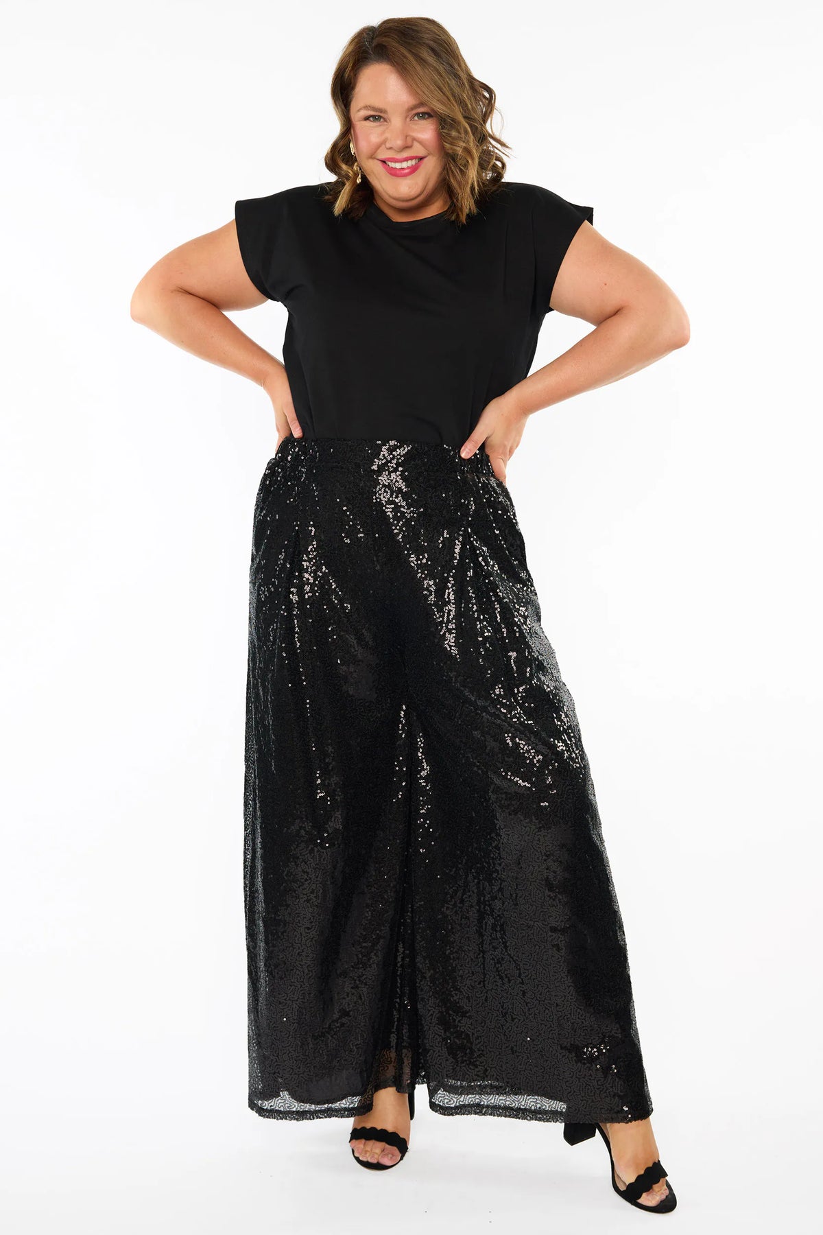 A woman modelling a black short sleeves top and a black sequin wide leg pant, standing against a plain white studio background with black heels.