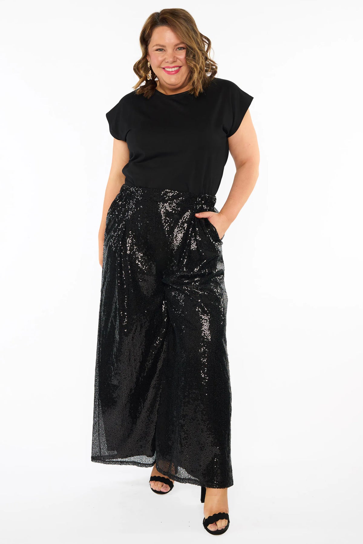 A woman modelling a black short sleeves top and a black sequin wide leg pant, standing against a plain white studio background with black heels.