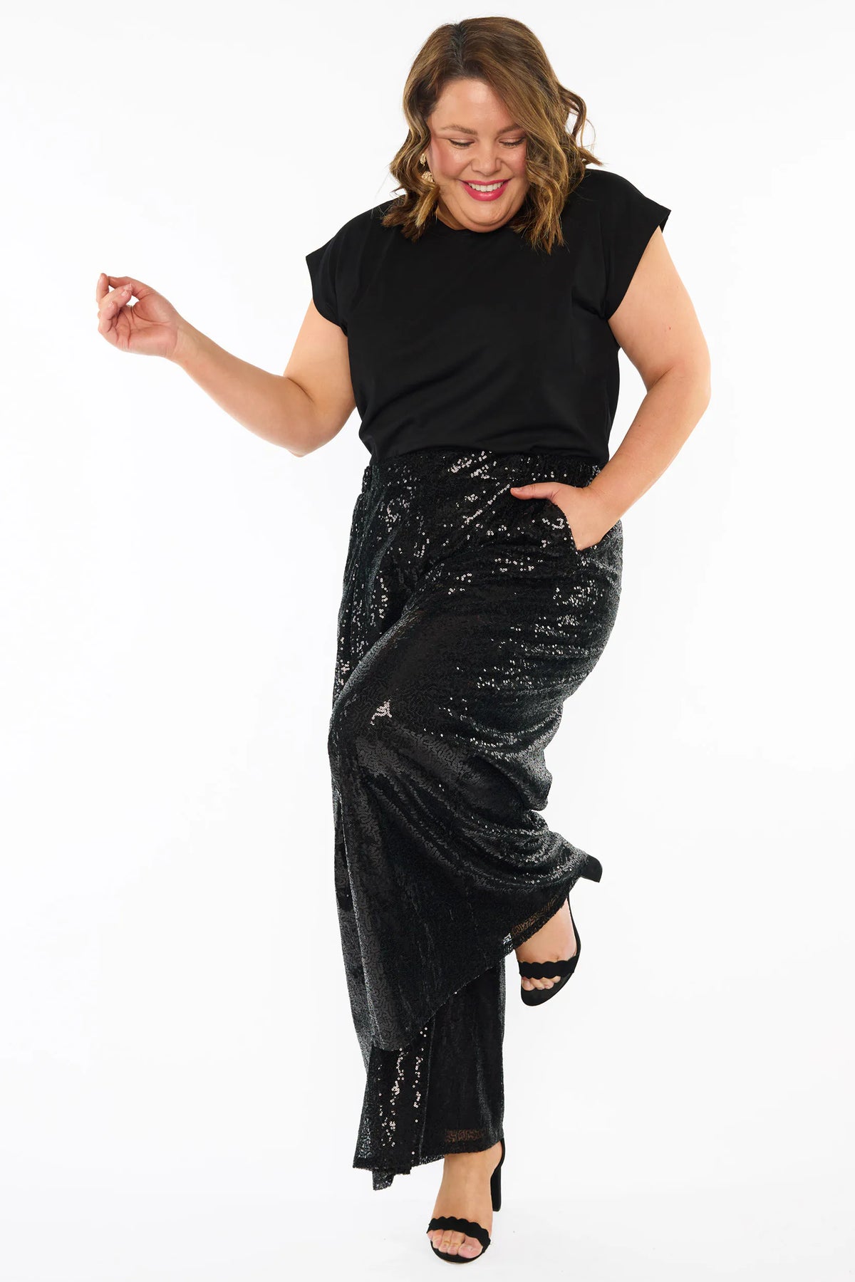 A woman modelling a black short sleeves top and a black sequin wide leg pant, standing against a plain white studio background with black heels.