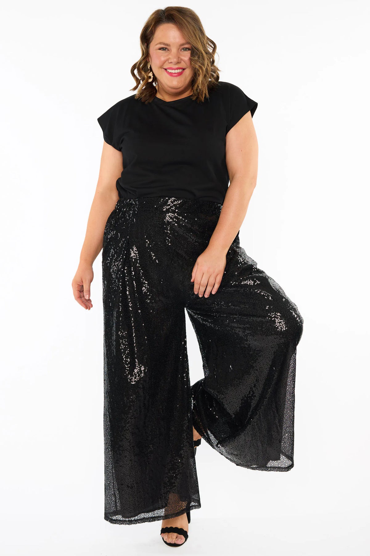 A woman modelling a black short sleeves top and a black sequin wide leg pant, standing against a plain white studio background with black heels.