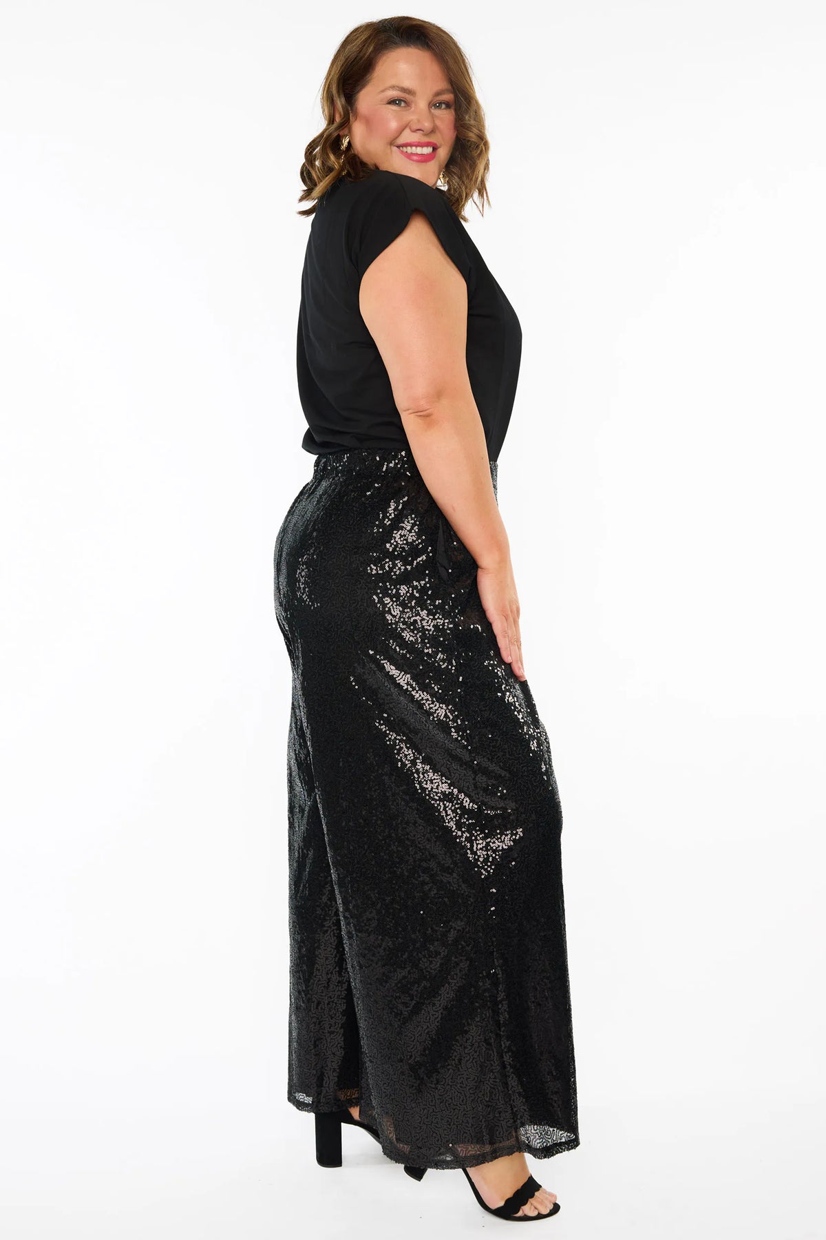 A woman modelling a black short sleeves top and a black sequin wide leg pant, standing against a plain white studio background with black heels.