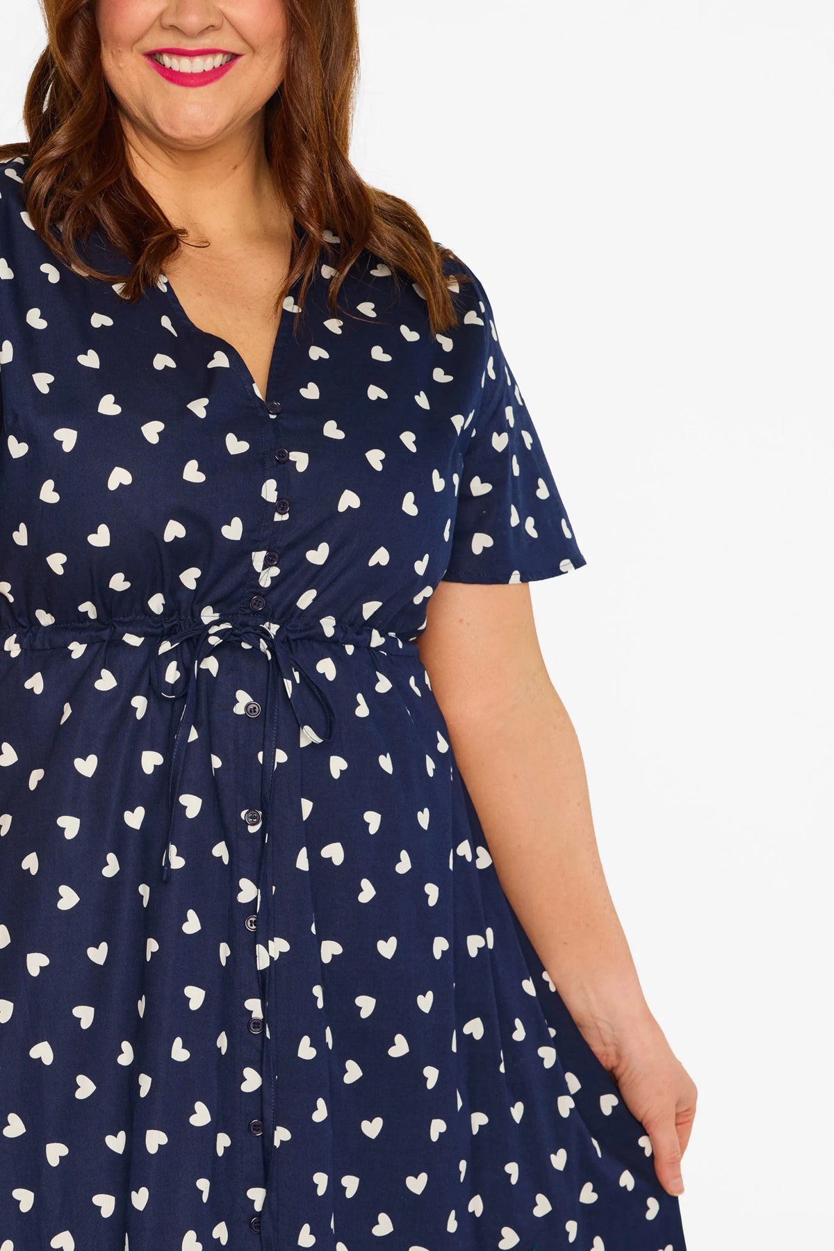 Close up of a woman modelling a navy blue midi dress with a white heart pattern, with V-neckline, short sleeves and an adjustable tie waist, standing against a plain white studio background.