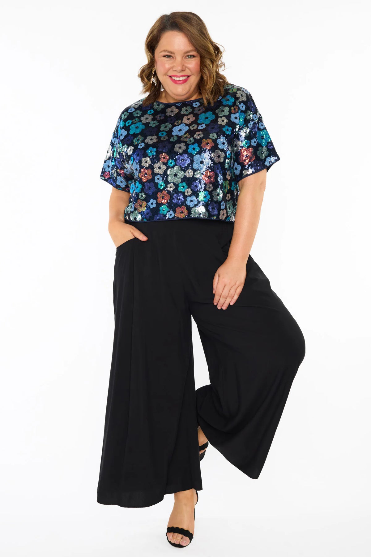 A woman modelling a short-sleeve top with a mix of blue tonal coloured sequin flowers on a black sequin background, paired with black wide-leg pants and black heeled sandals, against a plain white studio background.