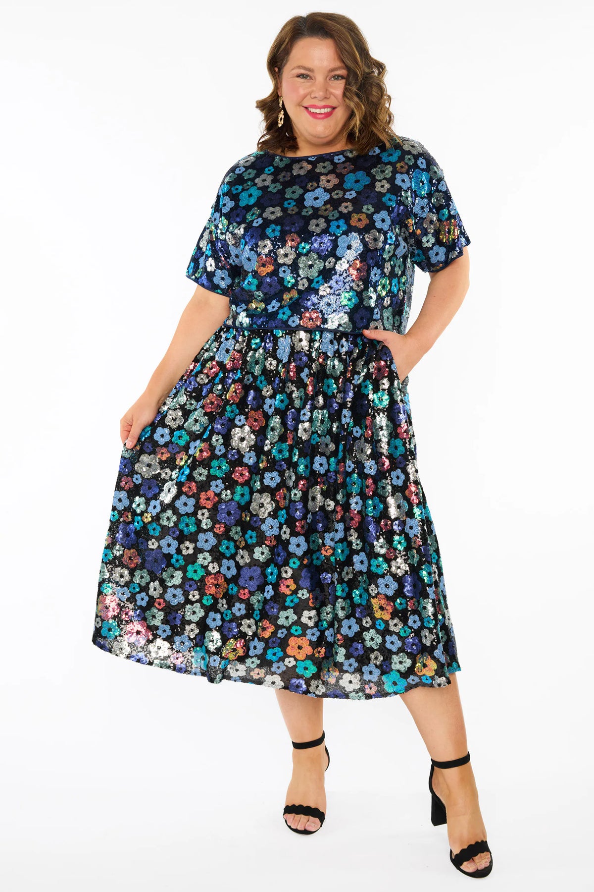 A woman modelling a sequins short-sleeve top with a mix of blue tonal coloured flowers on a black background, paired with a matching knee length skirt and black heels, against a plain white studio background.