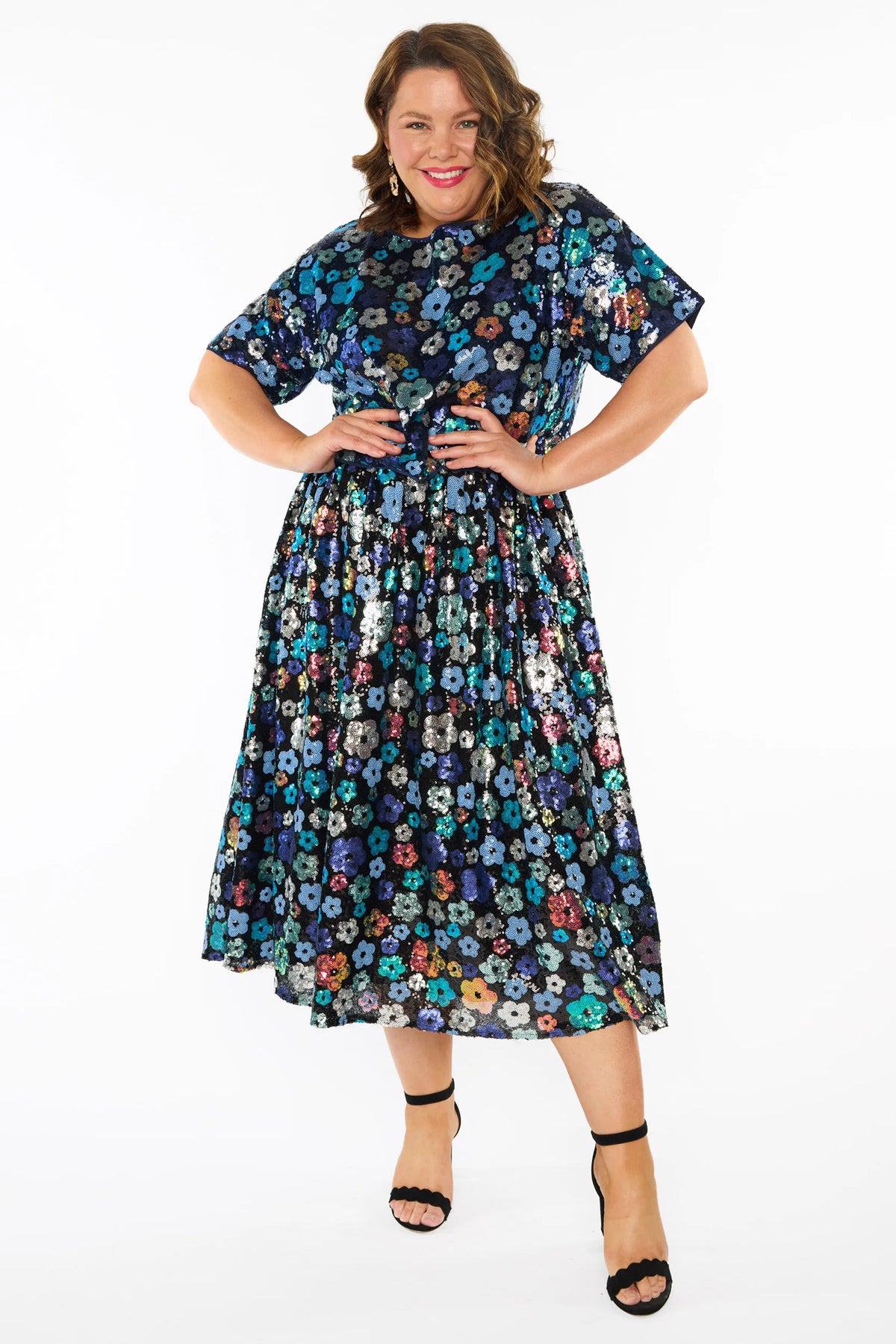 A woman modelling a sequins short-sleeve top with a mix of blue tonal coloured flowers on a black background, paired with a matching knee length skirt and black heels, against a plain white studio background.
