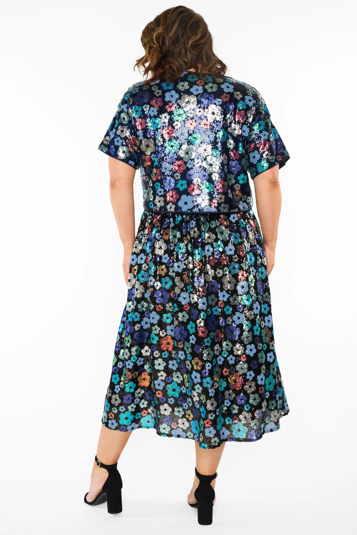 A woman modelling a sequins short-sleeve top with a mix of blue tonal coloured flowers on a black background, paired with a matching knee length skirt and black heels, against a plain white studio background.