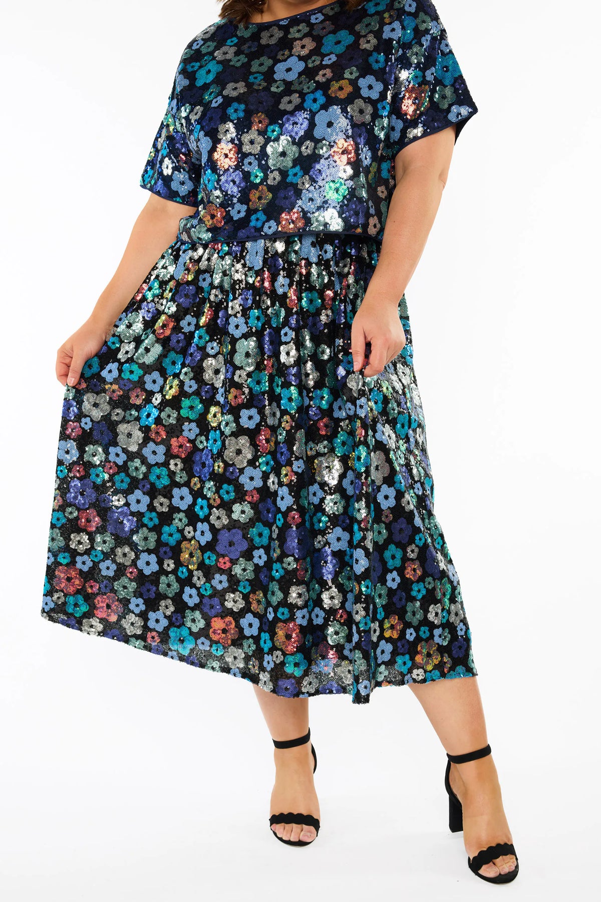 A woman modelling a sequins short-sleeve top with a mix of blue tonal coloured flowers on a black background, paired with a matching knee length skirt and black heels, against a plain white studio background.