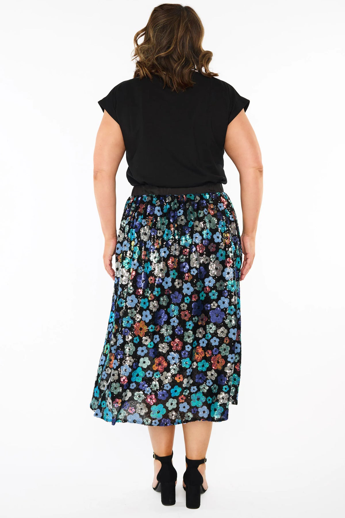 A woman modelling a short-sleeve black top, with a sequin knee length skirt featuring a mix of blue tonal coloured flowers on a black background. Paired with black heels, against a plain white studio background.