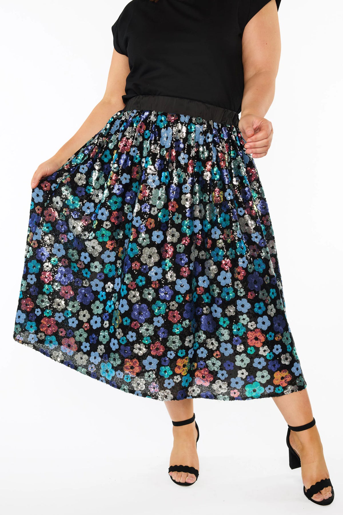 Close up of a woman modelling a short-sleeve black top, with a sequin knee length skirt featuring a mix of blue tonal coloured flowers on a black background. Paired with black heels, against a plain white studio background.