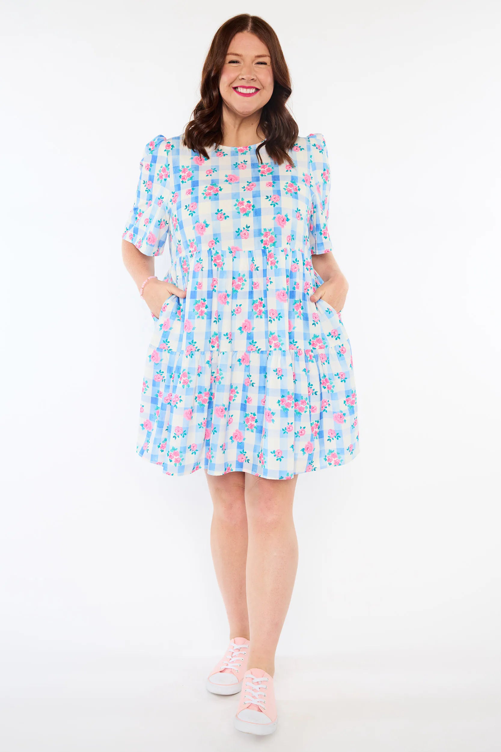 A woman modeling a short puff-sleeve dress with a blue, pink, and white floral gingham pattern, paired with light pink sneakers, against a plain white studio background.