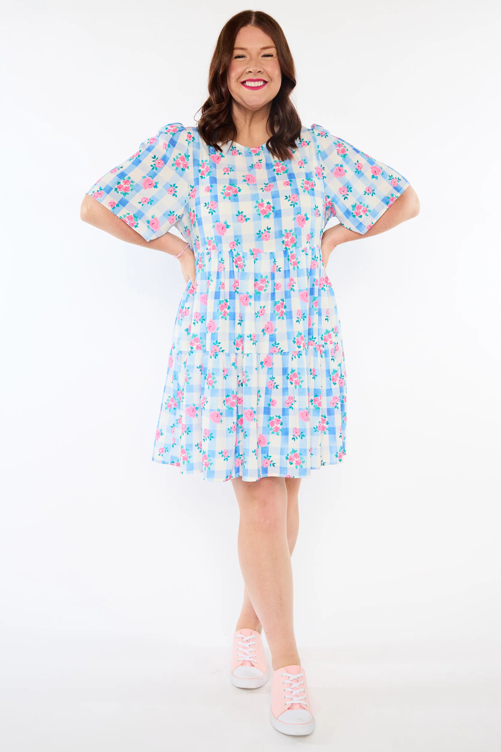 A woman modeling a short-sleeve tiered dress with a blue, pink, and white gingham pattern, paired with light pink sneakers, against a plain white studio background.