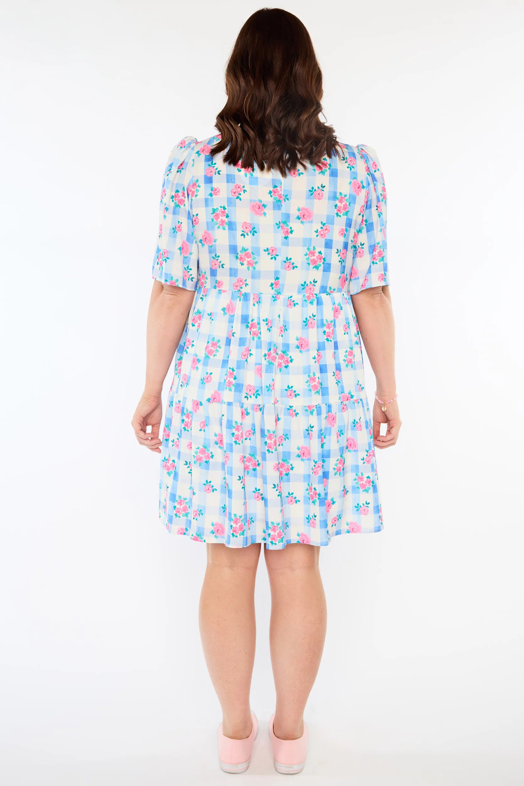 A woman viewed from the back wearing a short-sleeve tiered dress with a light blue, pink, and white floral print, paired with pale pink heels, against a plain white background.