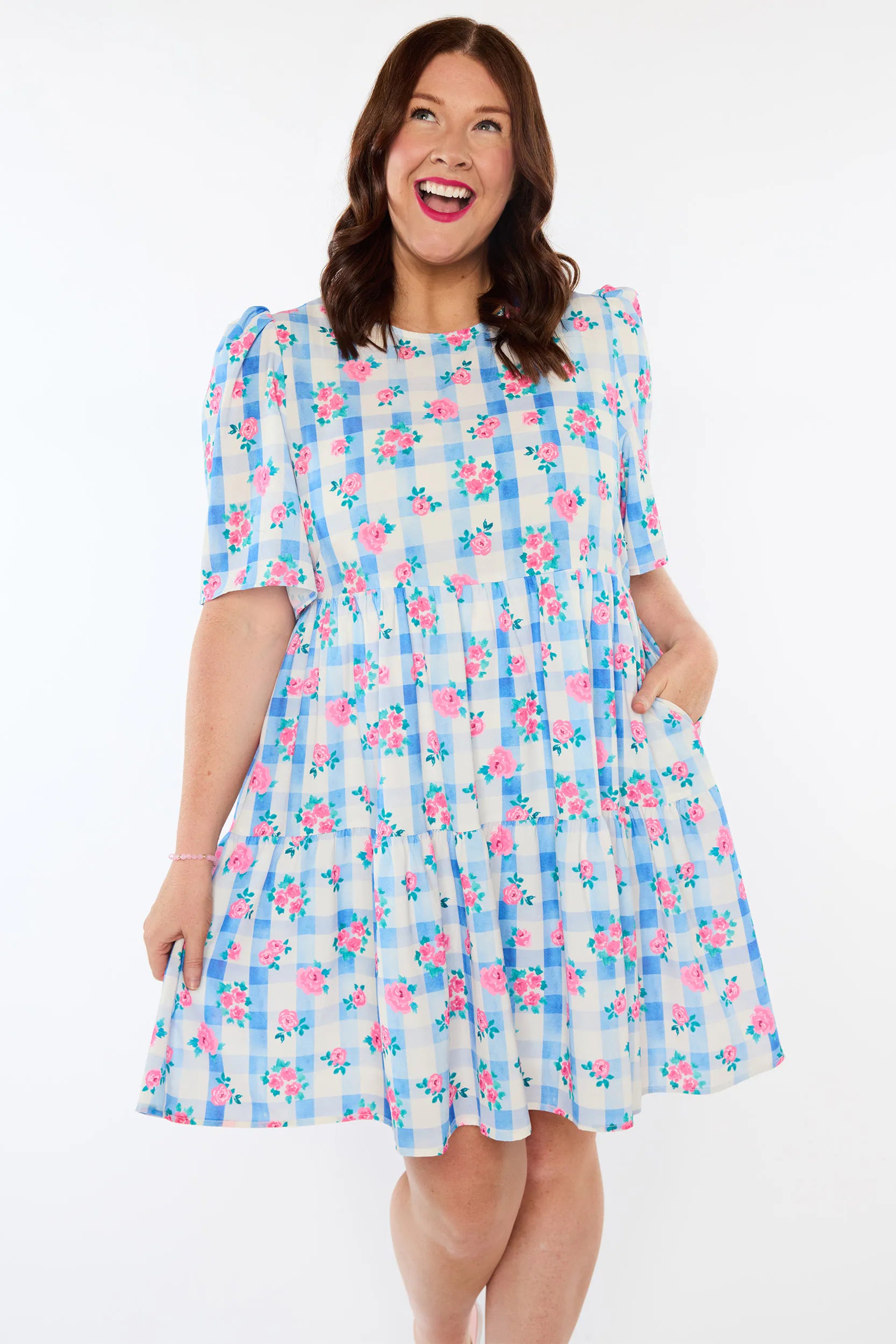 A woman modeling a short-sleeve tiered dress with a blue and white gingham pattern and pink floral print, set against a plain white background.