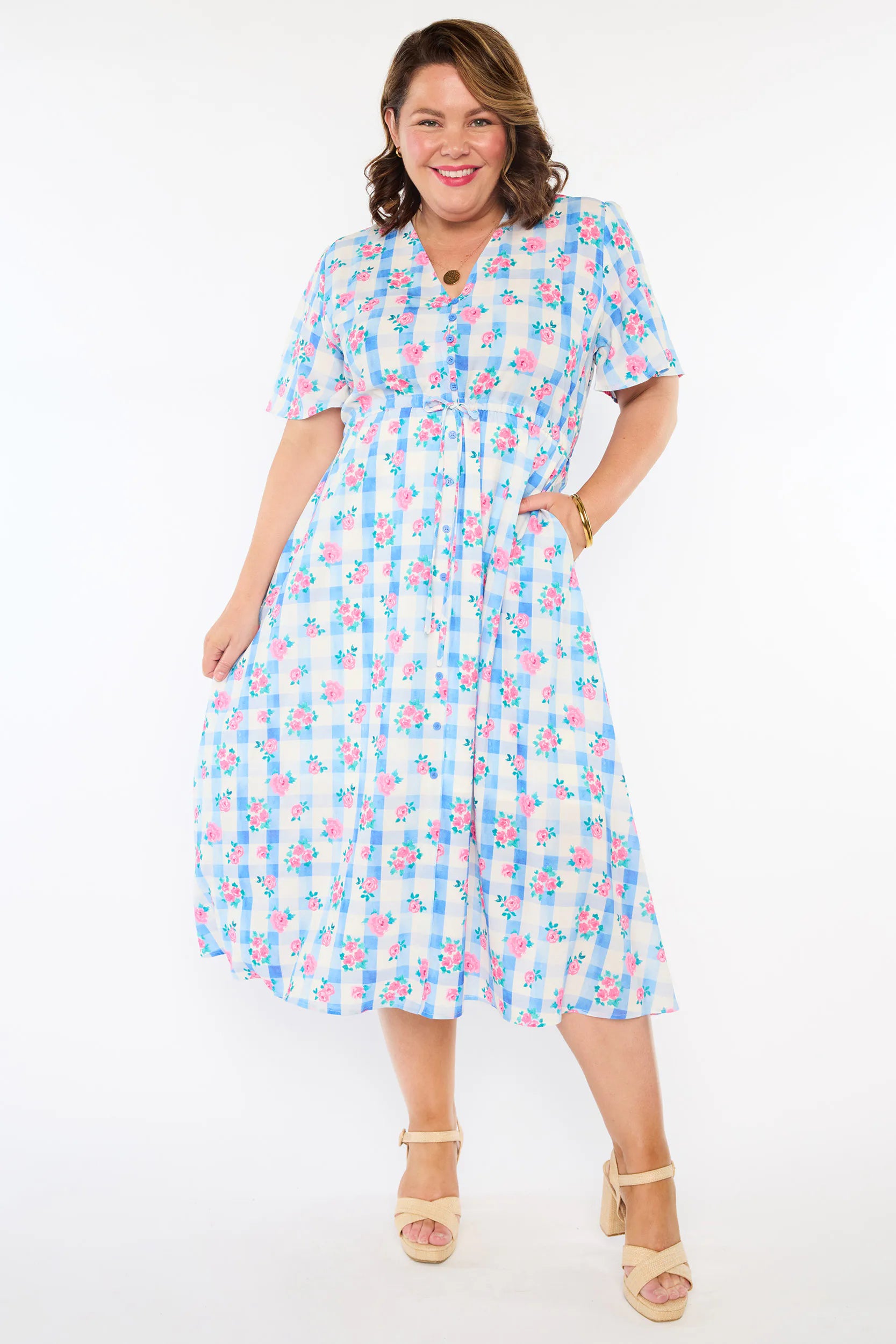 A woman modelling a short-sleeve, Midi-dress dress, with a button-up V-neckline & adjustable tie waist. A light blue and white gingham background covered in pink little roses, paired with cream heels, against a plain white studio background.