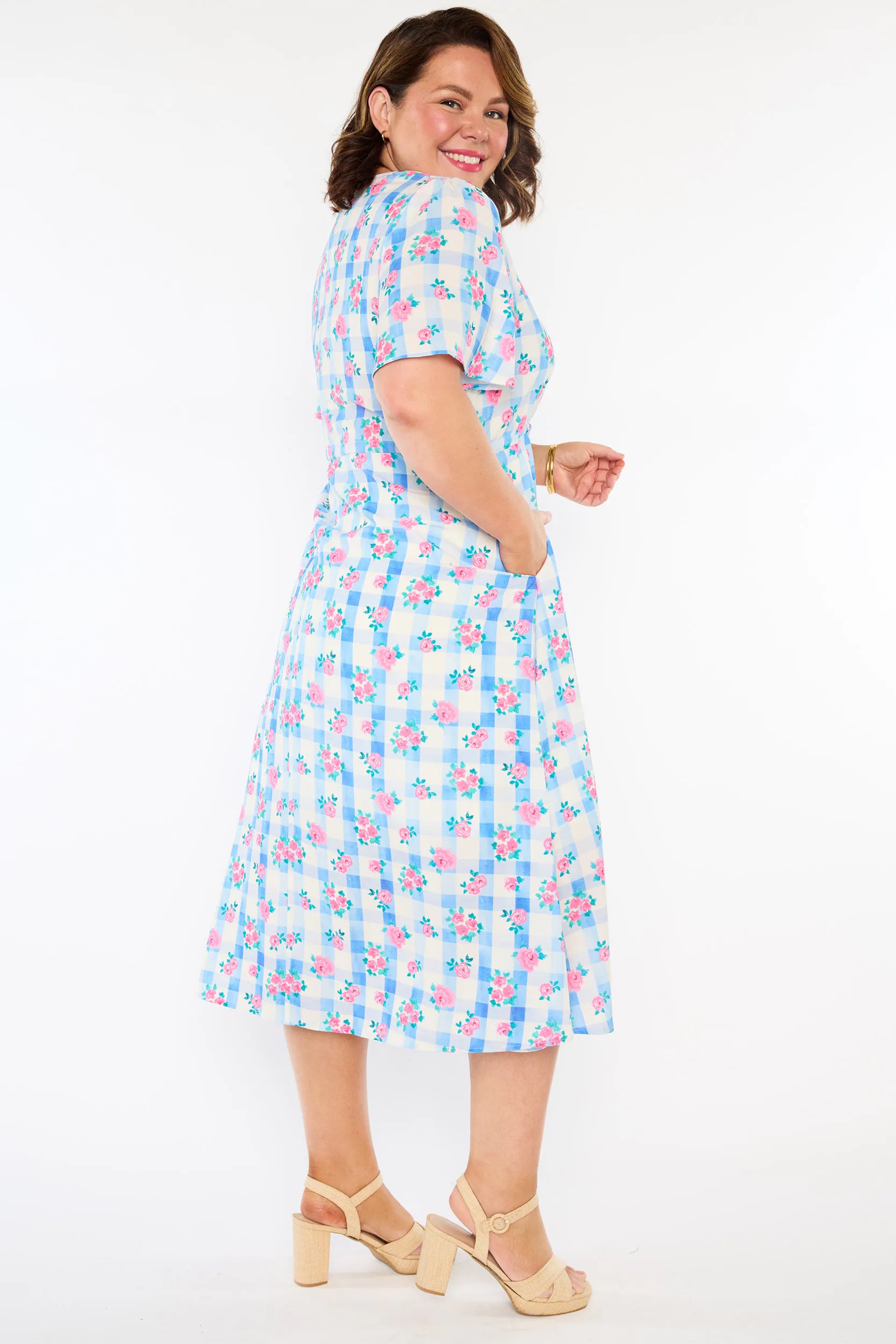 A woman modelling a short-sleeve, Midi-dress dress, with a button-up V-neckline & adjustable tie waist. A light blue and white gingham background covered in pink little roses, paired with cream heels, against a plain white studio background.
