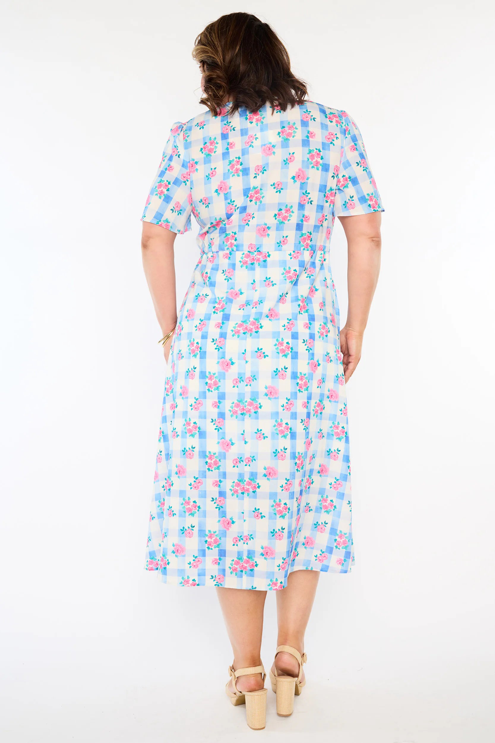A woman modelling a short-sleeve, Midi-dress dress, with a button-up V-neckline & adjustable tie waist. A light blue and white gingham background covered in pink little roses, paired with cream heels, against a plain white studio background.