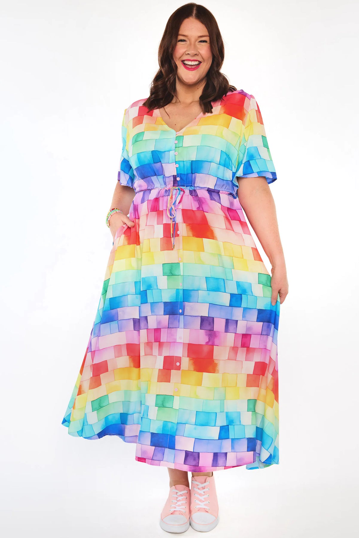 A woman modeling a short-sleeve V-neck midi dress with a colorful pixelated block pattern in shades of blue, green, yellow, orange, pink, purple, and red, paired with light pink sneakers against a white studio background.