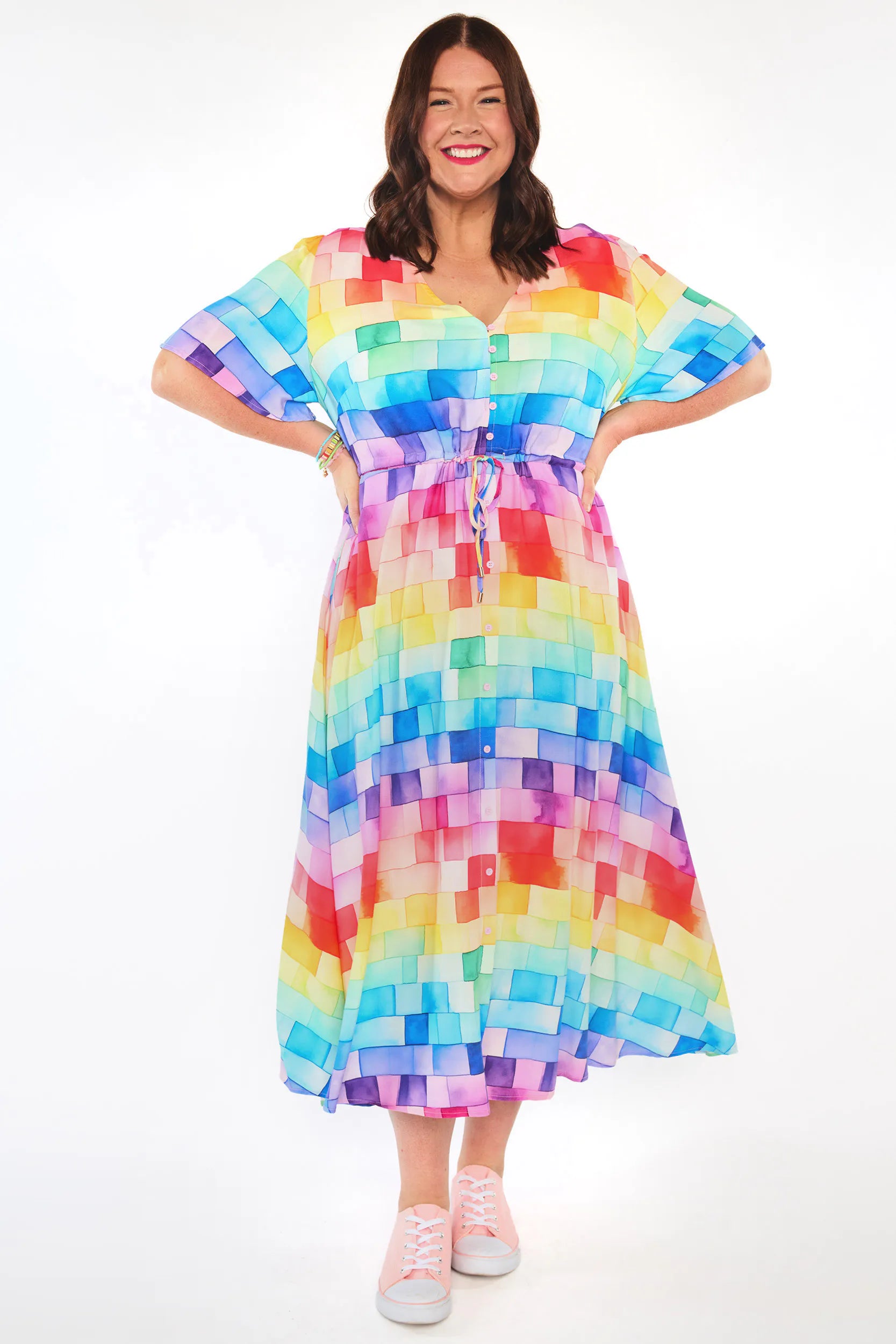 A woman modeling a short-sleeved midi dress with a colorful rainbow pixelated block pattern, standing against a plain white studio background.