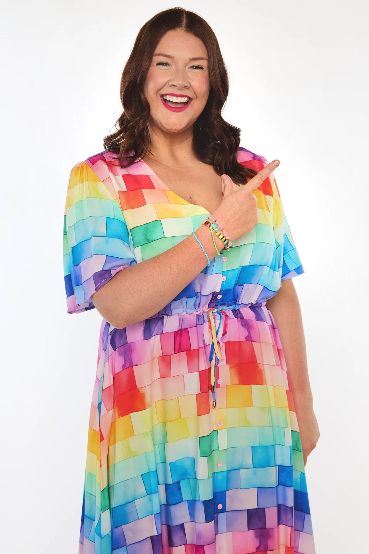 A woman smiling and pointing at a colorful dress with a rainbow block pattern, set against a plain white background.