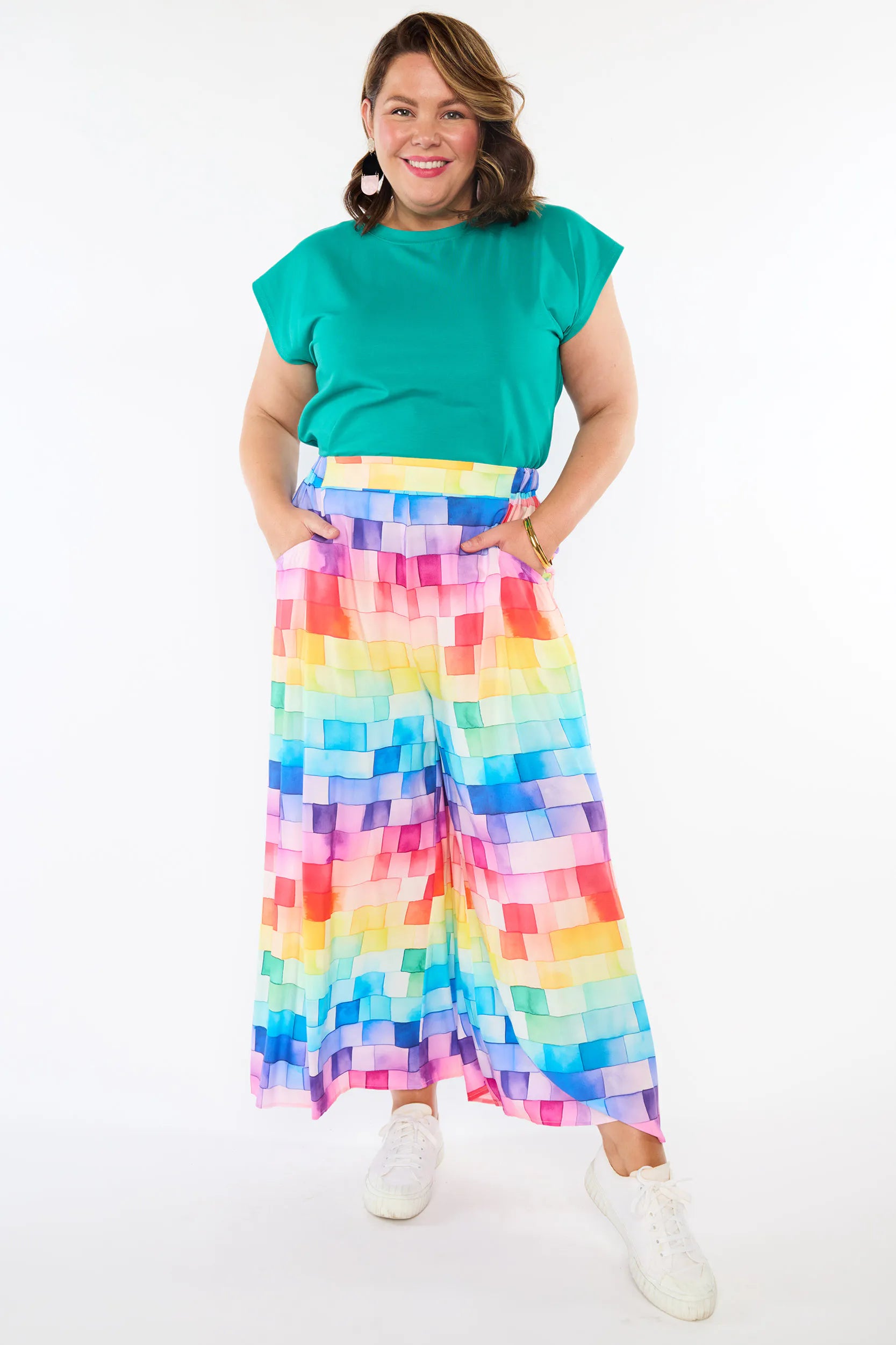 A woman modeling a vibrant rainbow pixelated print wide-leg skirt paired with a bright green short-sleeve top and white sneakers, set against a plain white studio background.