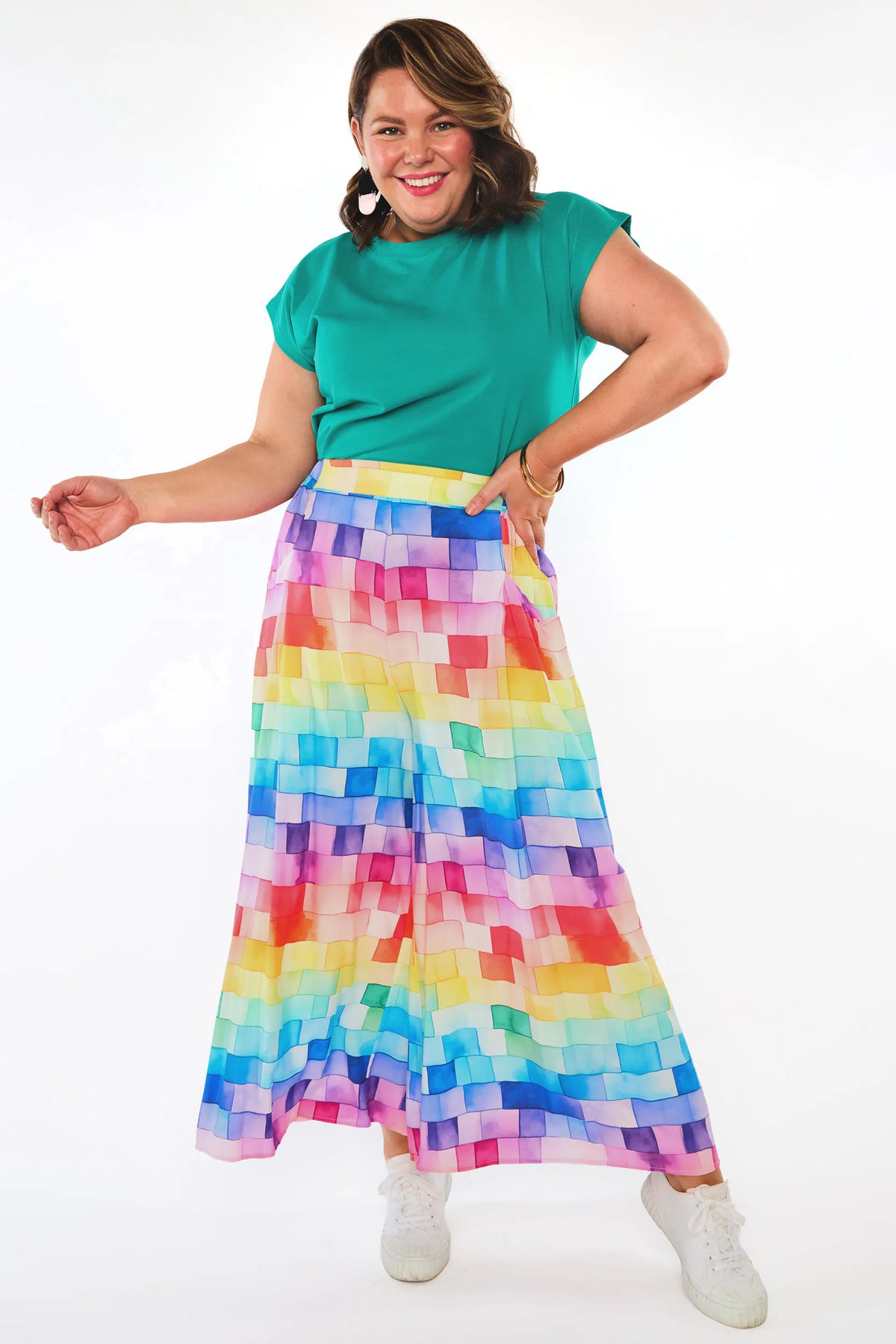 A woman modeling a vibrant, multicolor pixelated-patterned maxi skirt paired with a solid teal green short-sleeve top and white sneakers, set against a plain white studio background.