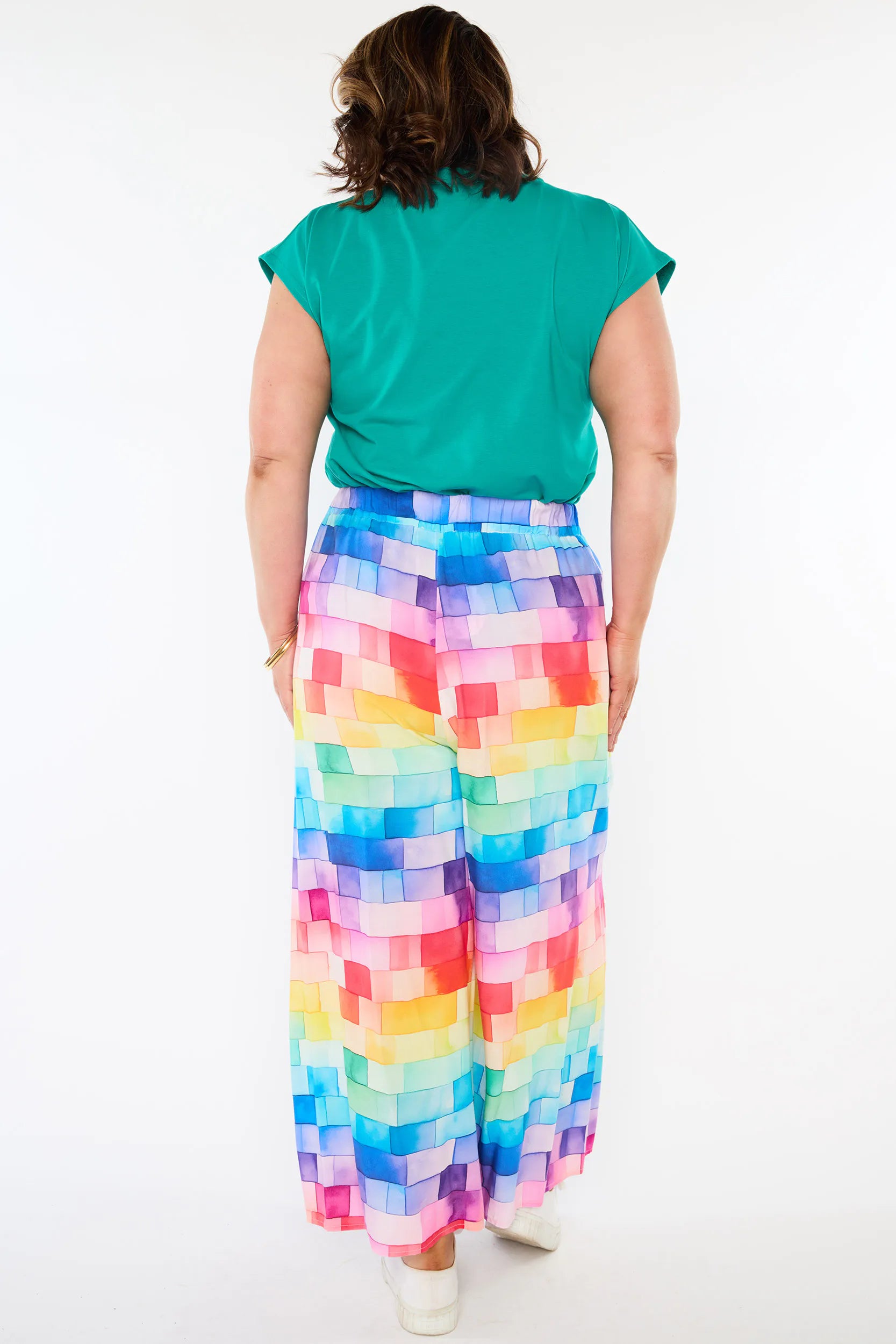 A woman viewed from behind wearing a bright green short-sleeve top and high-waisted, wide-leg pants with a colorful pixelated rainbow pattern, set against a plain white studio background.