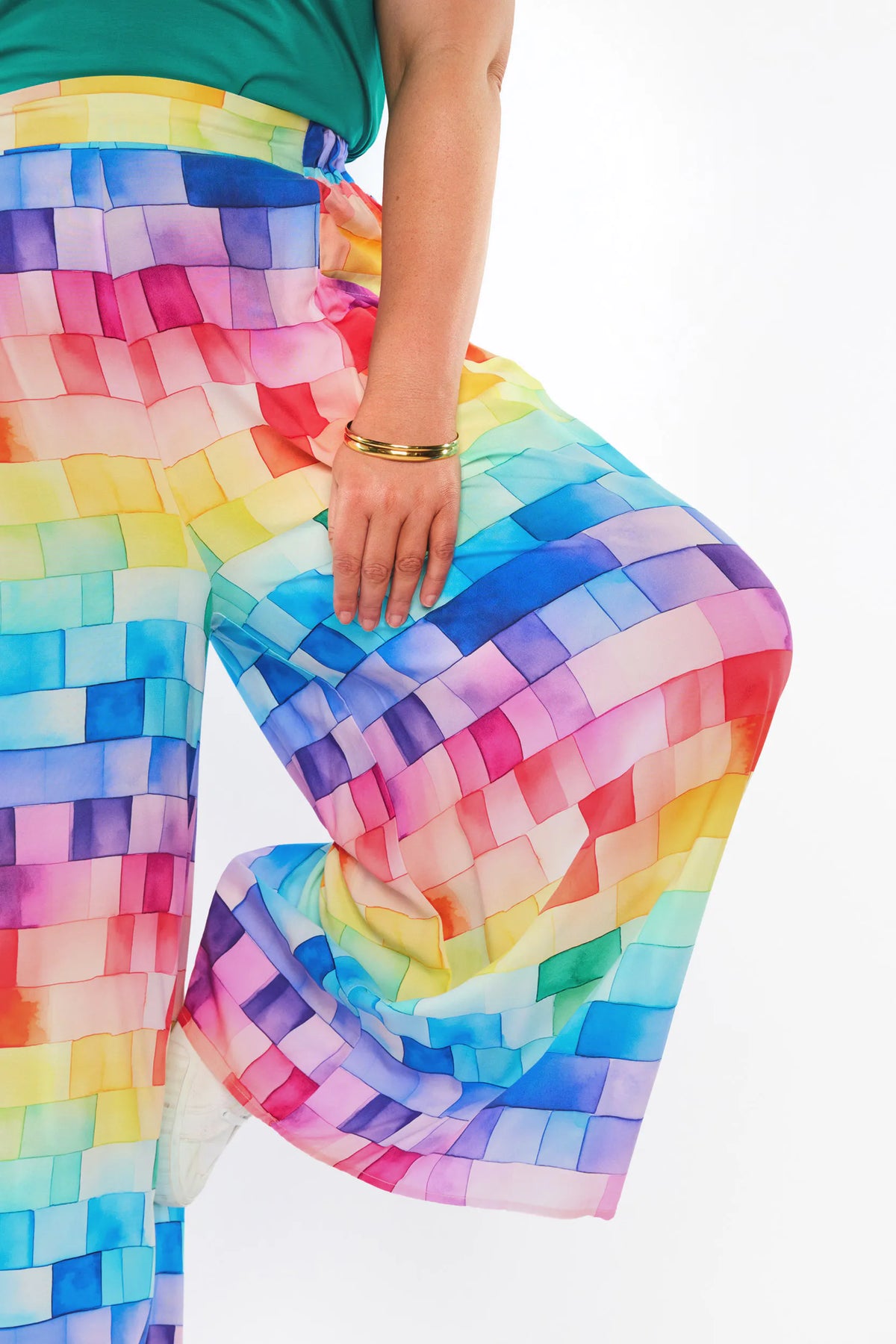 A close-up of a person wearing wide-leg pants with a colorful rainbow checkered pattern, paired with a green top. The person is shown from the waist down, with one leg bent, against a plain white background.