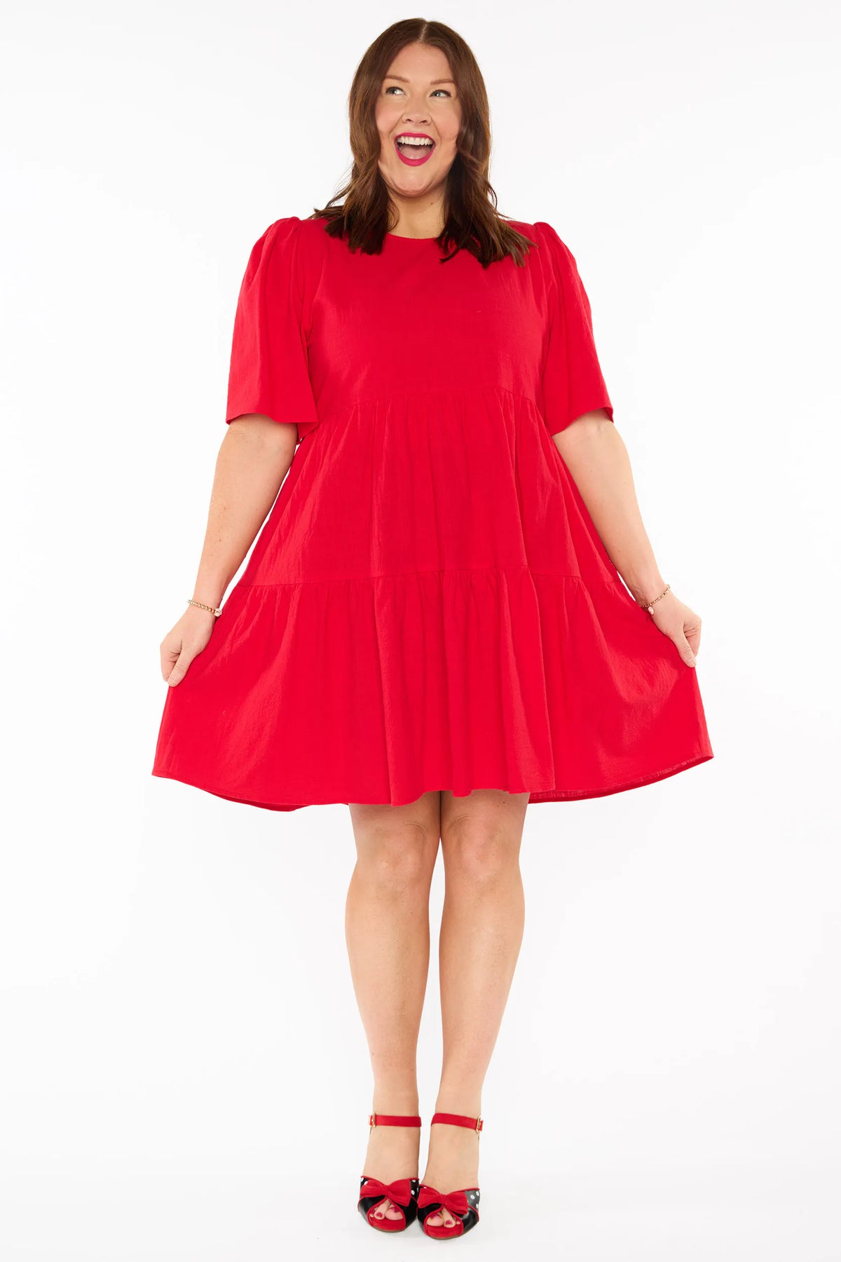 A woman modelling a bright red tiered mini dress with short puff sleeves and red bow heels against a plain white studio background.