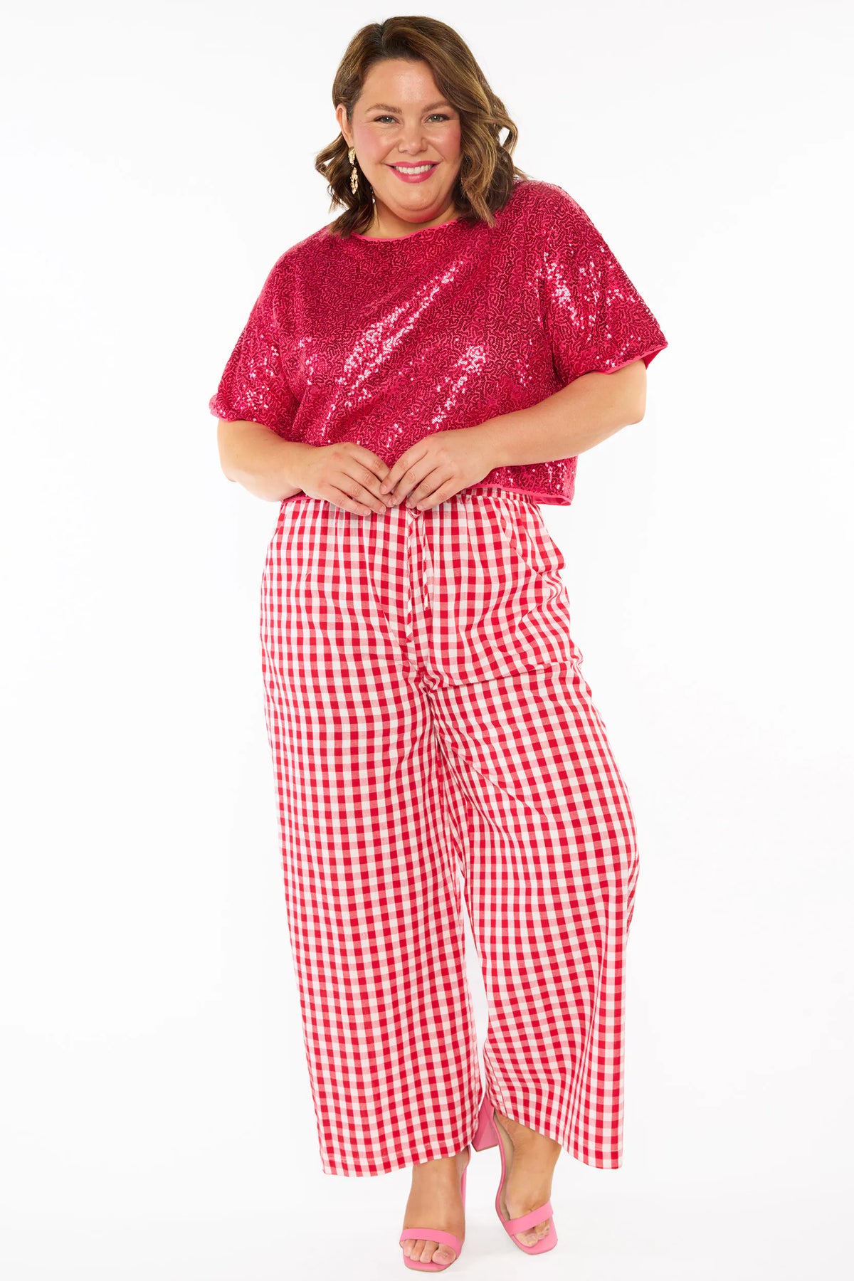 A woman modelling a two-piece outfit consisting of a bright pink sequins short-sleeve top and red-and-white gingham wide-leg pants, paired with pink heels, against a plain white background.