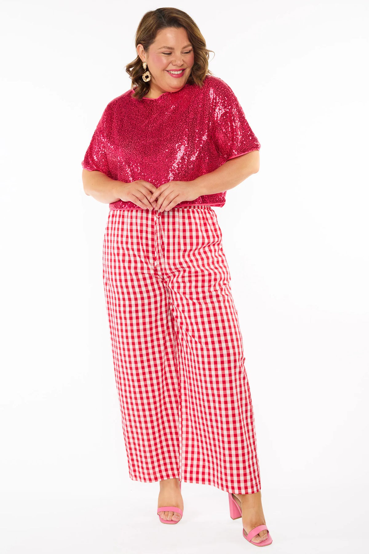 A woman modelling a two-piece outfit consisting of a bright pink sequins short-sleeve top and red-and-white gingham wide-leg pants, paired with pink heels, against a plain white background.