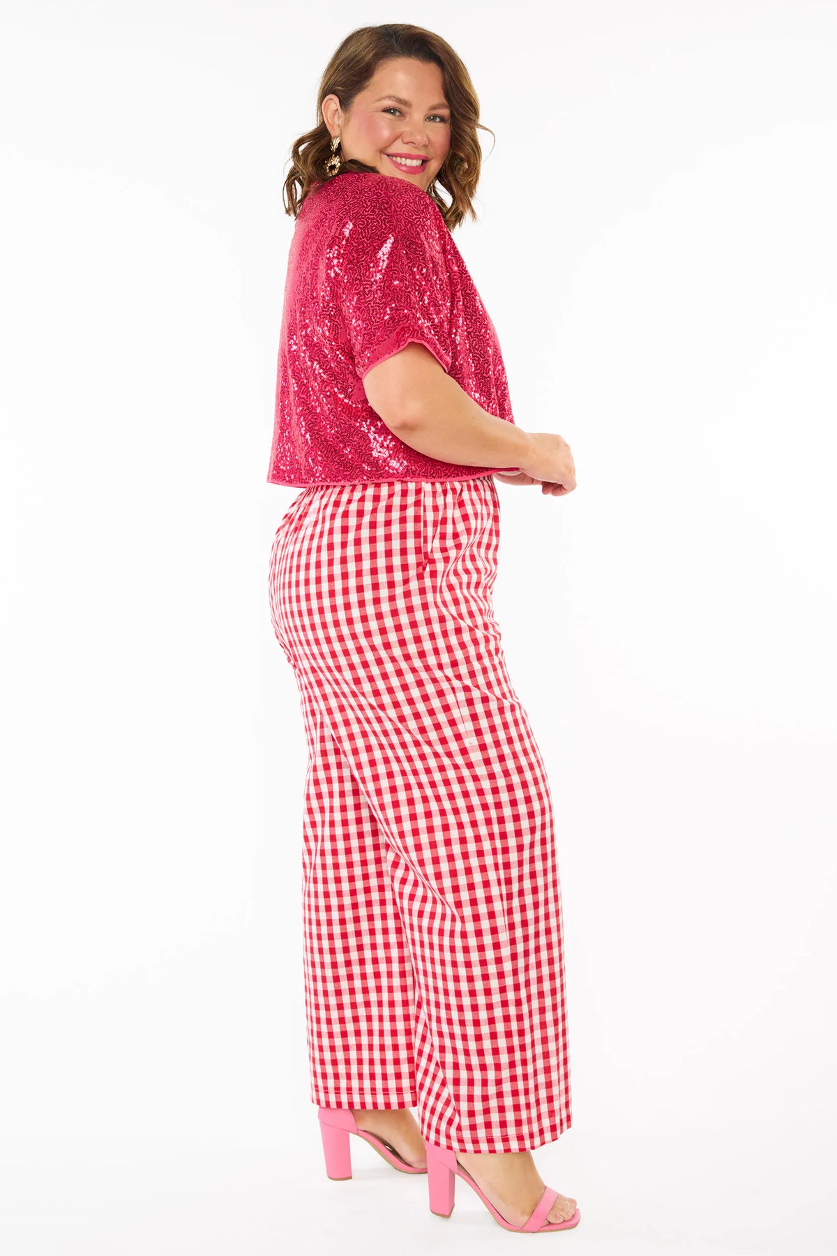 A woman modelling on the side a two-piece outfit consisting of a bright pink sequins short-sleeve top and red-and-white gingham wide-leg pants, paired with pink heels, against a plain white background.