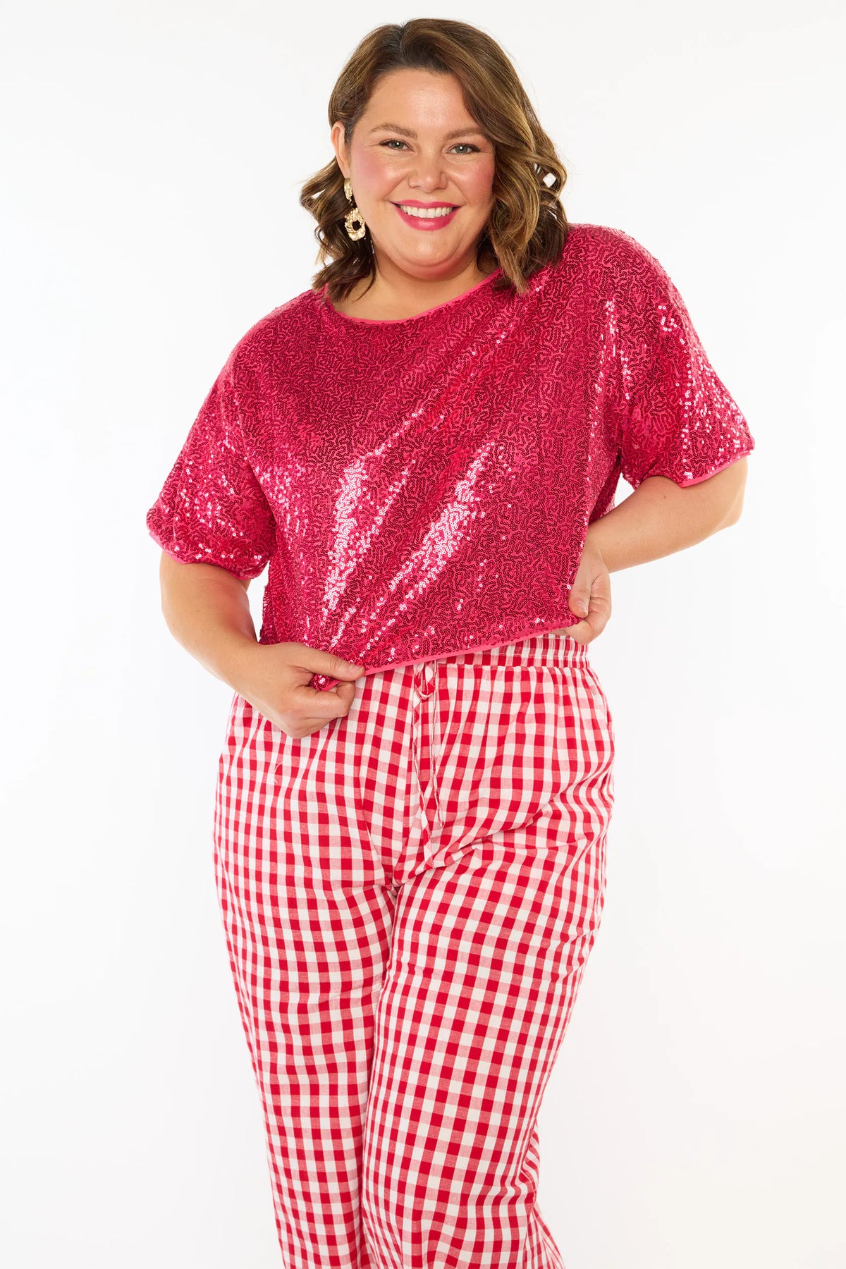 A woman modelling a two-piece outfit consisting of a bright pink sequins short-sleeve top and red-and-white gingham wide-leg pants, paired with pink heels, against a plain white background.