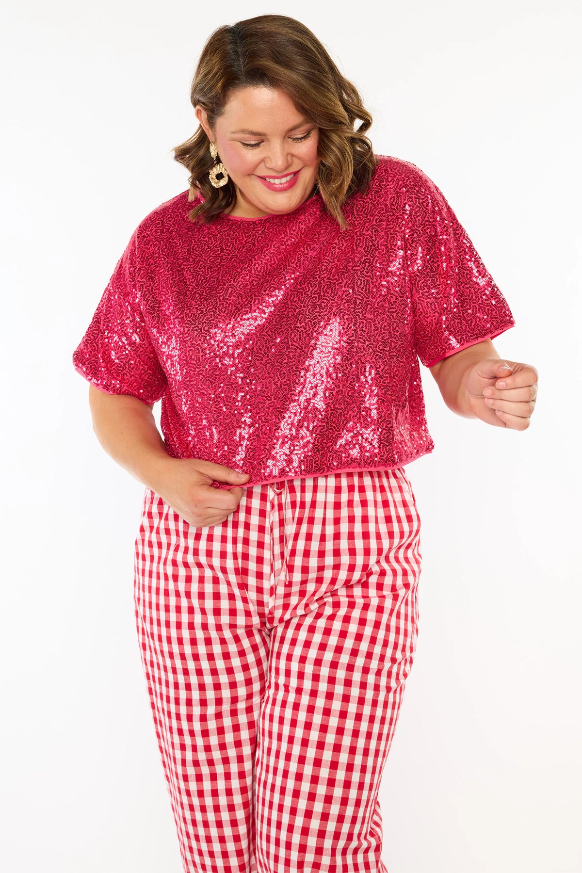 A woman modelling a two-piece outfit consisting of a bright pink sequins short-sleeve top and red-and-white gingham wide-leg pants, paired with pink heels, against a plain white background.