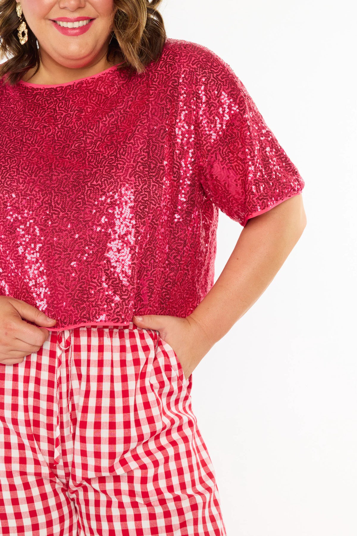A close ups of a woman modelling a two-piece outfit consisting of a bright pink sequins short-sleeve top and red-and-white gingham wide-leg pants, paired with pink heels, against a plain white background.