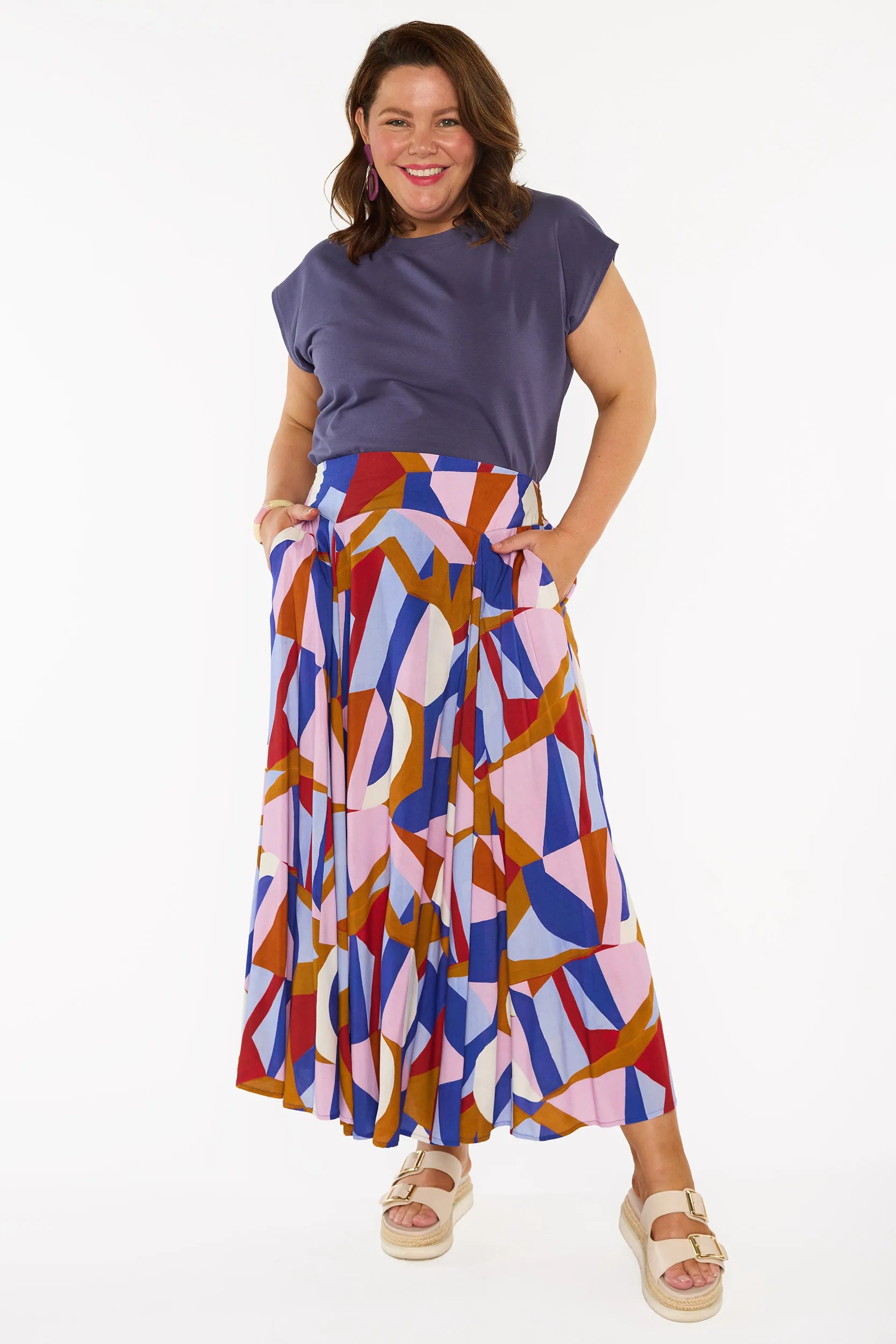 A woman modelling a two-piece outfit featuring a slate purple short-sleeve top and a high-waisted, pleated midi skirt with a colourful geometric abstract print in pink, blue, red, and brown, paired with beige platform sandals against a white studio background.