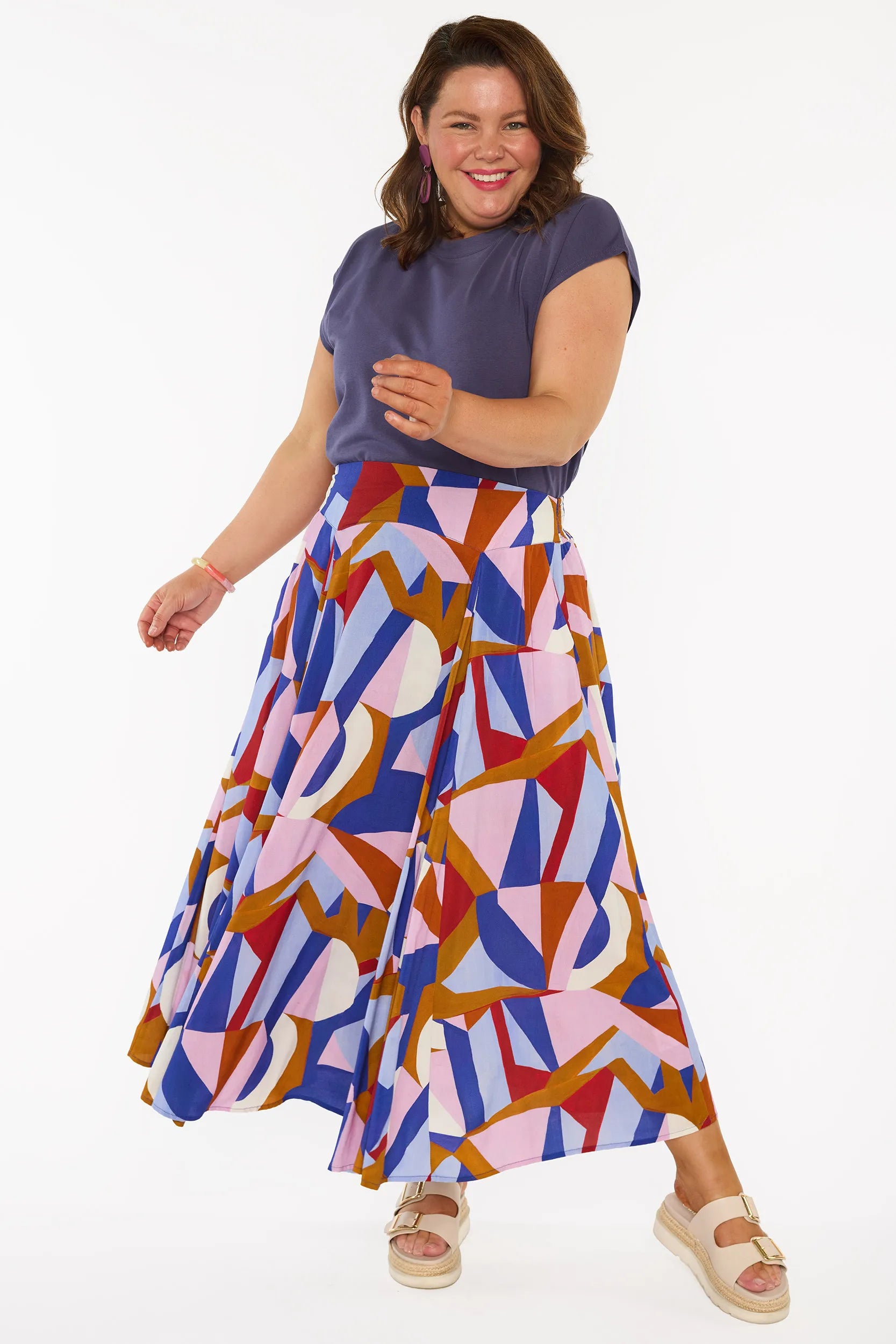 A woman modeling a sleeveless, high-waisted maxi skirt with a colorful geometric abstract print in blue, pink, red, and brown, paired with a solid navy blue top and beige platform sandals, against a plain white studio background.