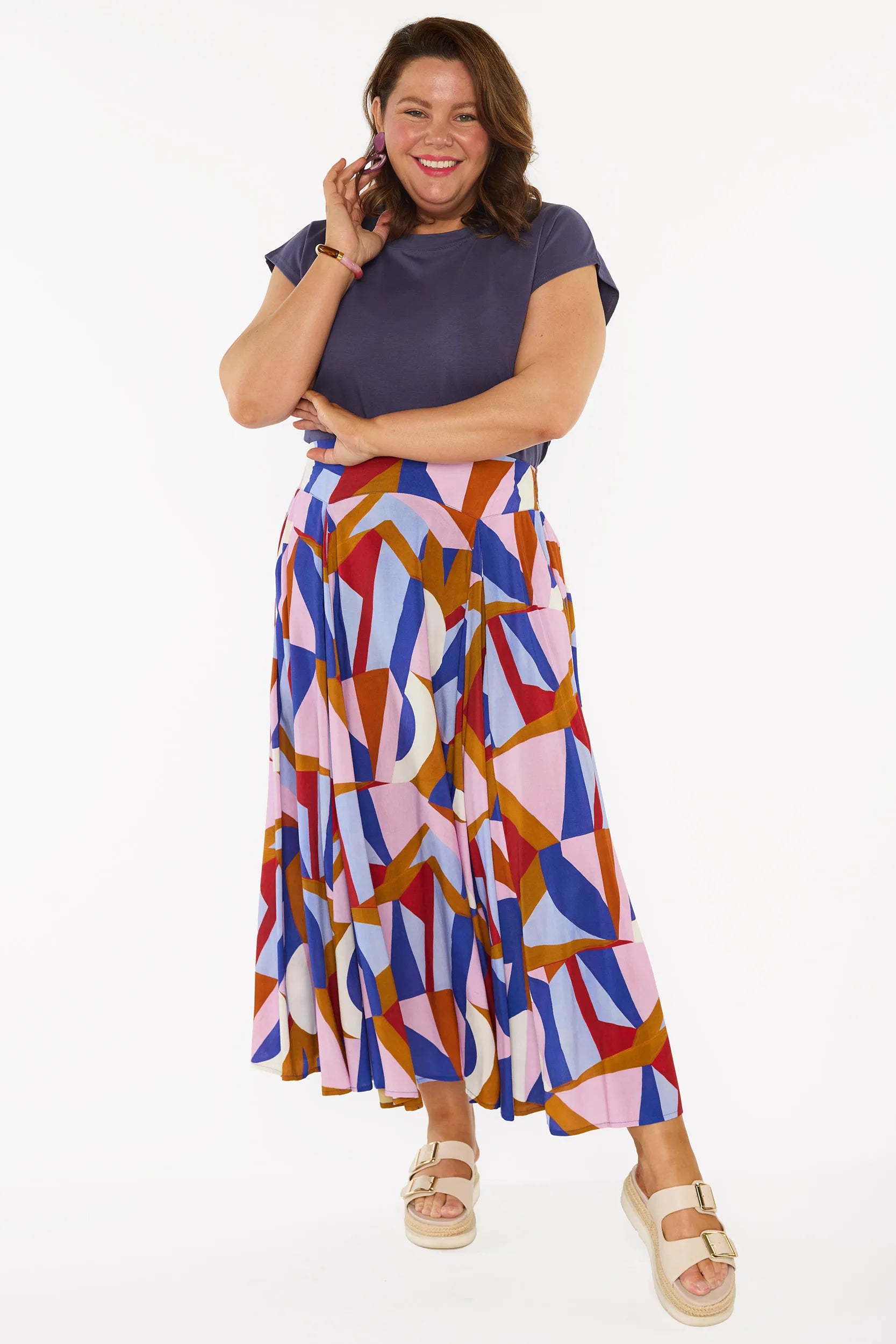 A woman modelling a sleeveless, high-waisted maxi skirt with a colourful geometric abstract print in blue, pink, red, and brown, paired with a solid navy blue top and beige platform sandals, against a plain white studio background.