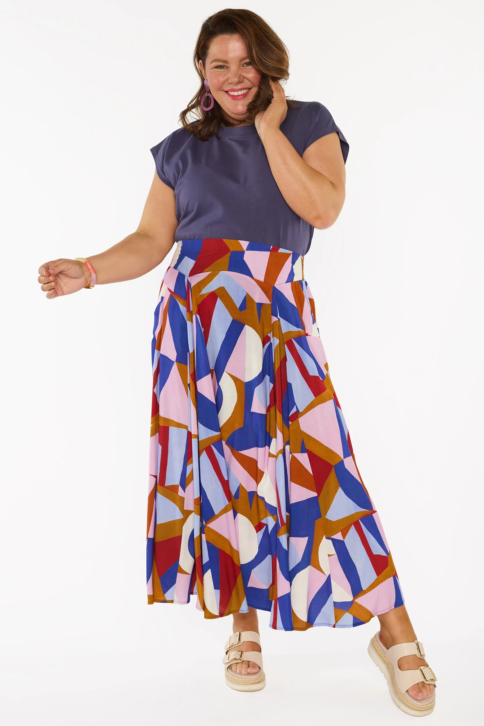 A woman modelling a sleeveless, high-waisted maxi skirt with a colourful geometric abstract print in blue, pink, red, and brown, paired with a solid navy blue top and beige platform sandals, against a plain white studio background.