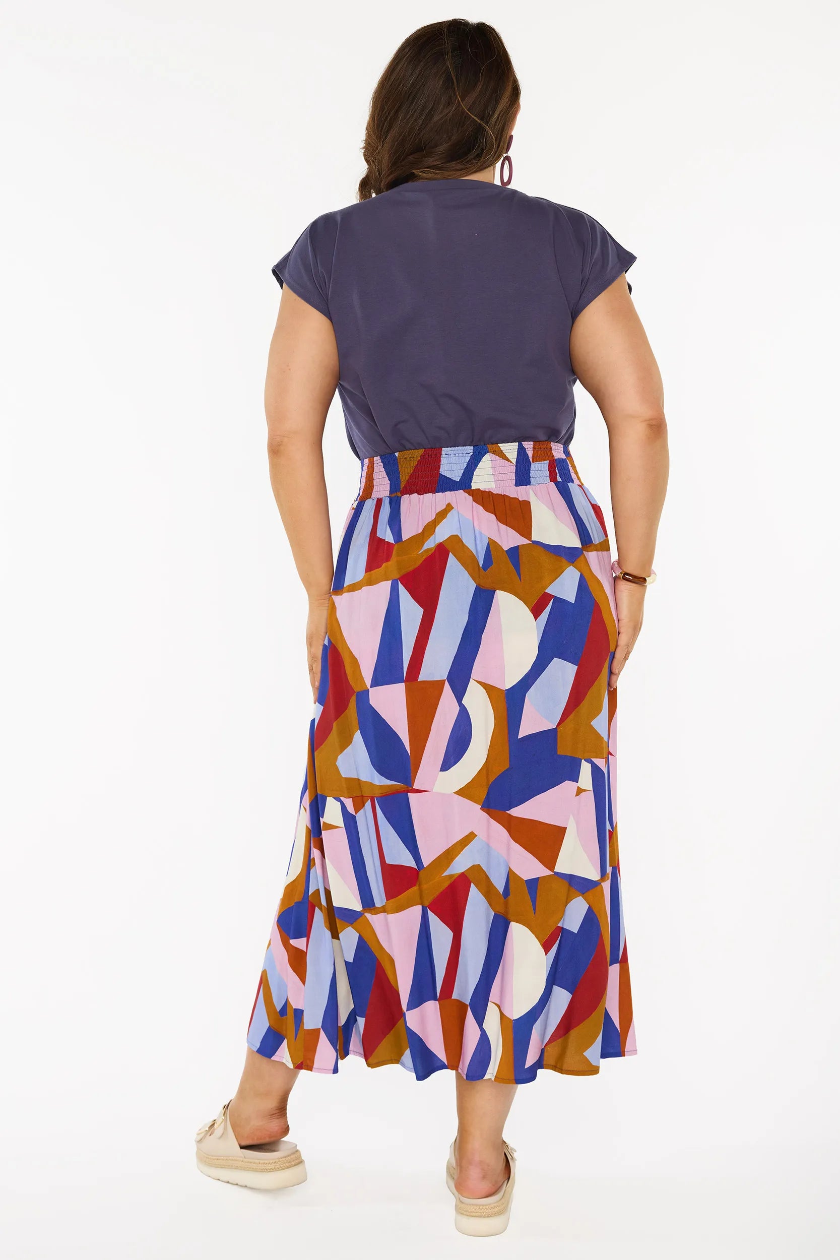 A woman modelling a sleeveless, high-waisted maxi skirt with a colourful geometric abstract print in blue, pink, red, and brown, paired with a solid navy blue top and beige platform sandals, against a plain white studio background.