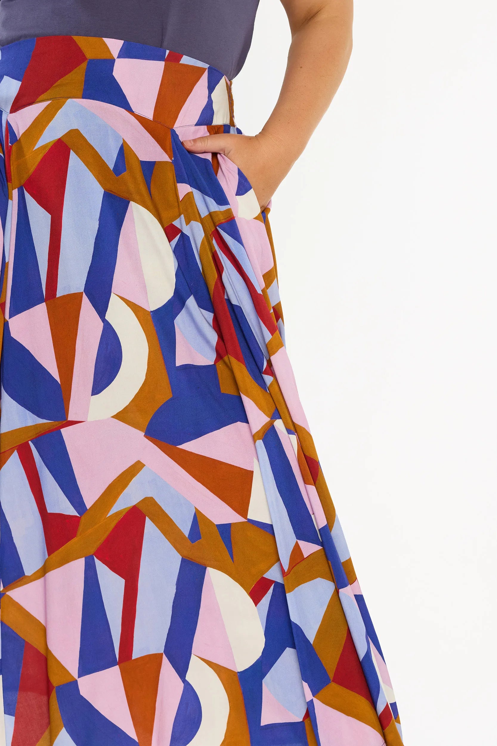 A woman modelling a sleeveless, high-waisted maxi skirt with a colourful geometric abstract print in blue, pink, red, and brown, paired with a solid navy blue top and beige platform sandals, against a plain white studio background.